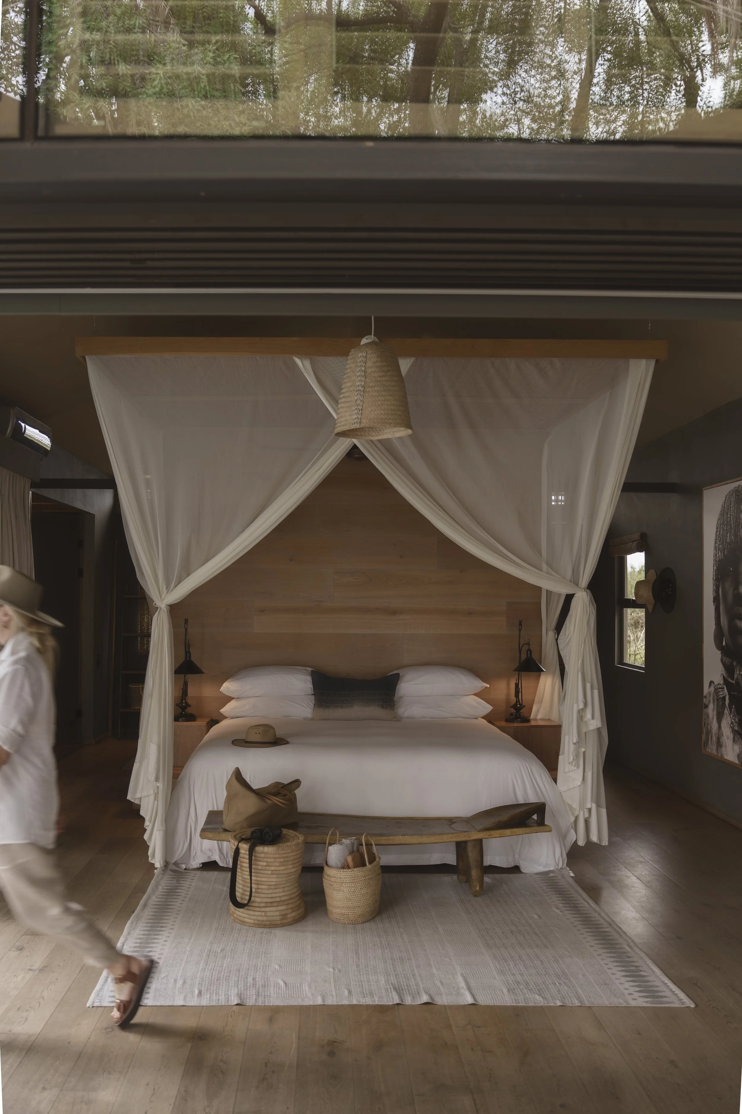 A bedroom with a large bed covered in white linens and four pillows, framed by a white sheer canopy, with a wooden headboard and two black wall-mounted lamps. The room has a wooden floor, a white rug, and some baskets and a hat on a bench at the foot of the bed. A soft, natural light illuminates the room, with windows on two sides and a ceiling light fixture with a woven shade hanging above the bed.