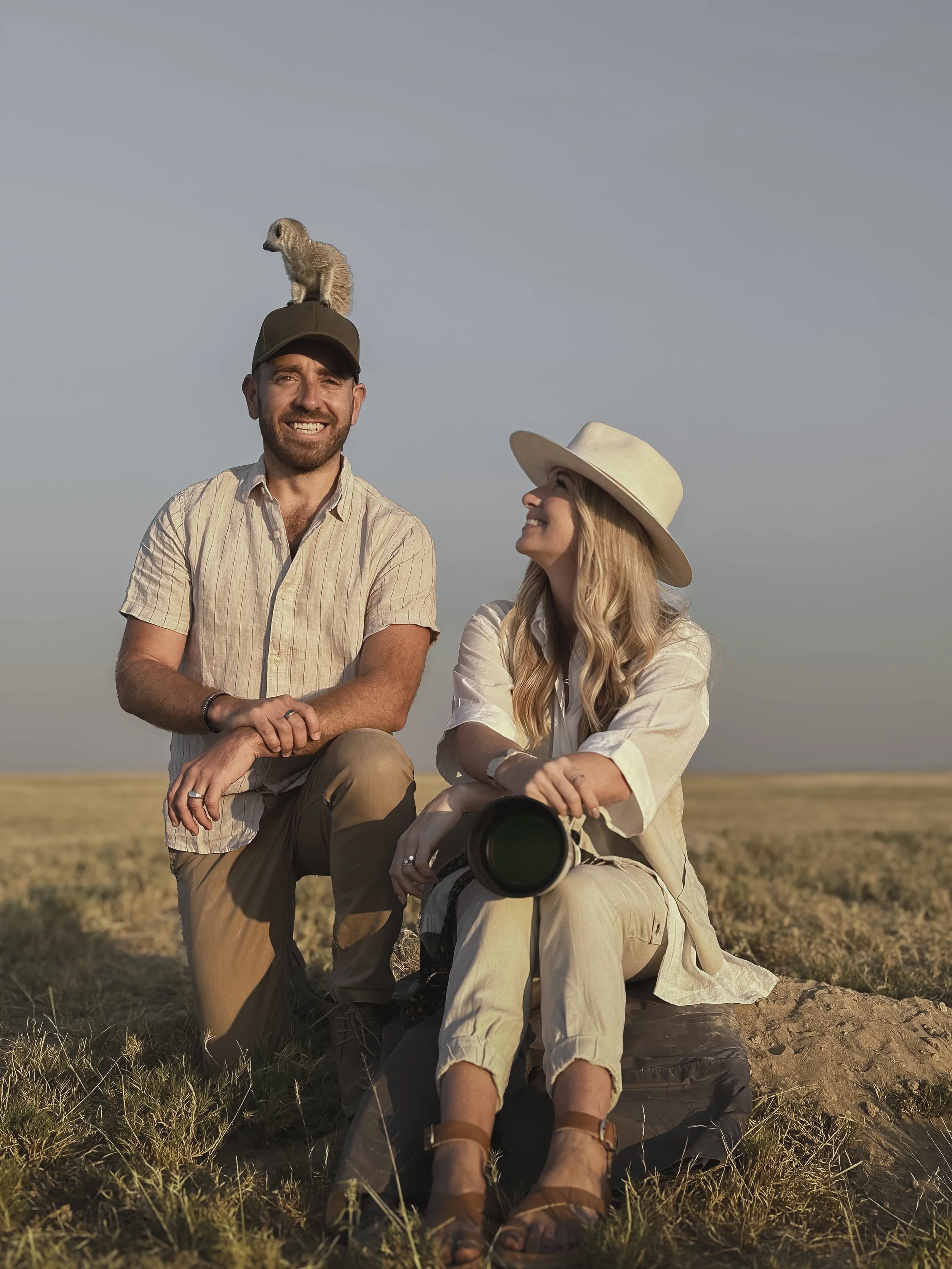 Jemma Park and her husband Guy sitting in the grass with a meerkat perched on his cap