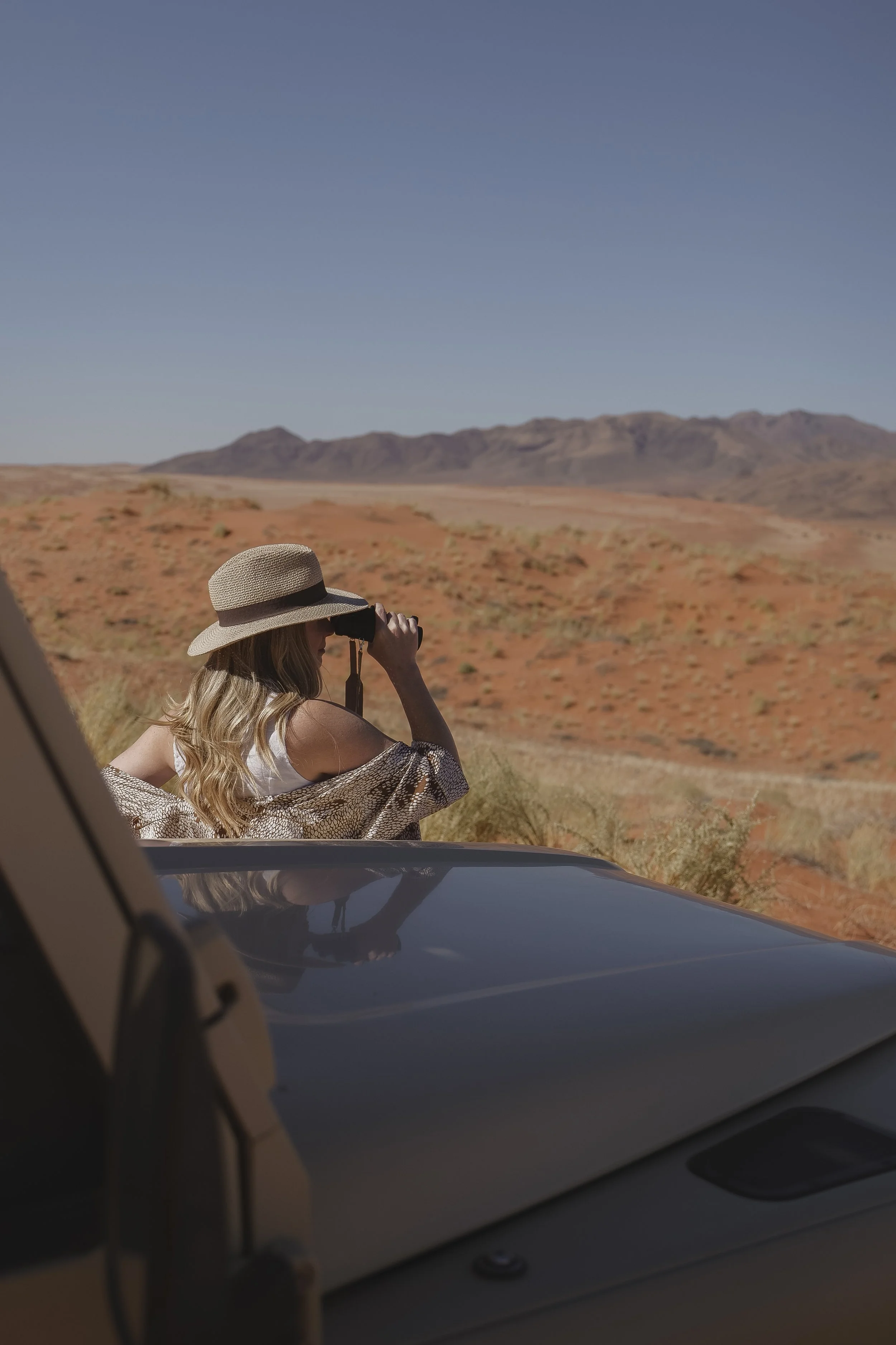 Woman with long blonde hair wearing a wide-brimmed hat and sunglasses, sitting by a car window in a desert landscape with mountains in the background, taking a photo or looking through binoculars.