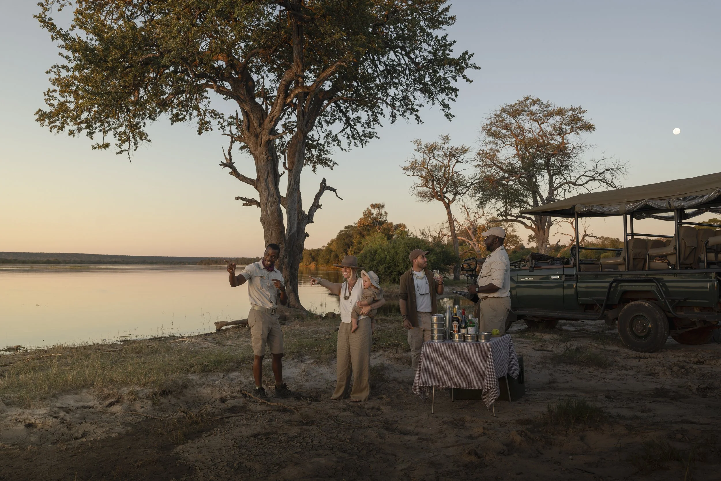 Family and safari guides gathered for a sundowner stop beside a river at dusk