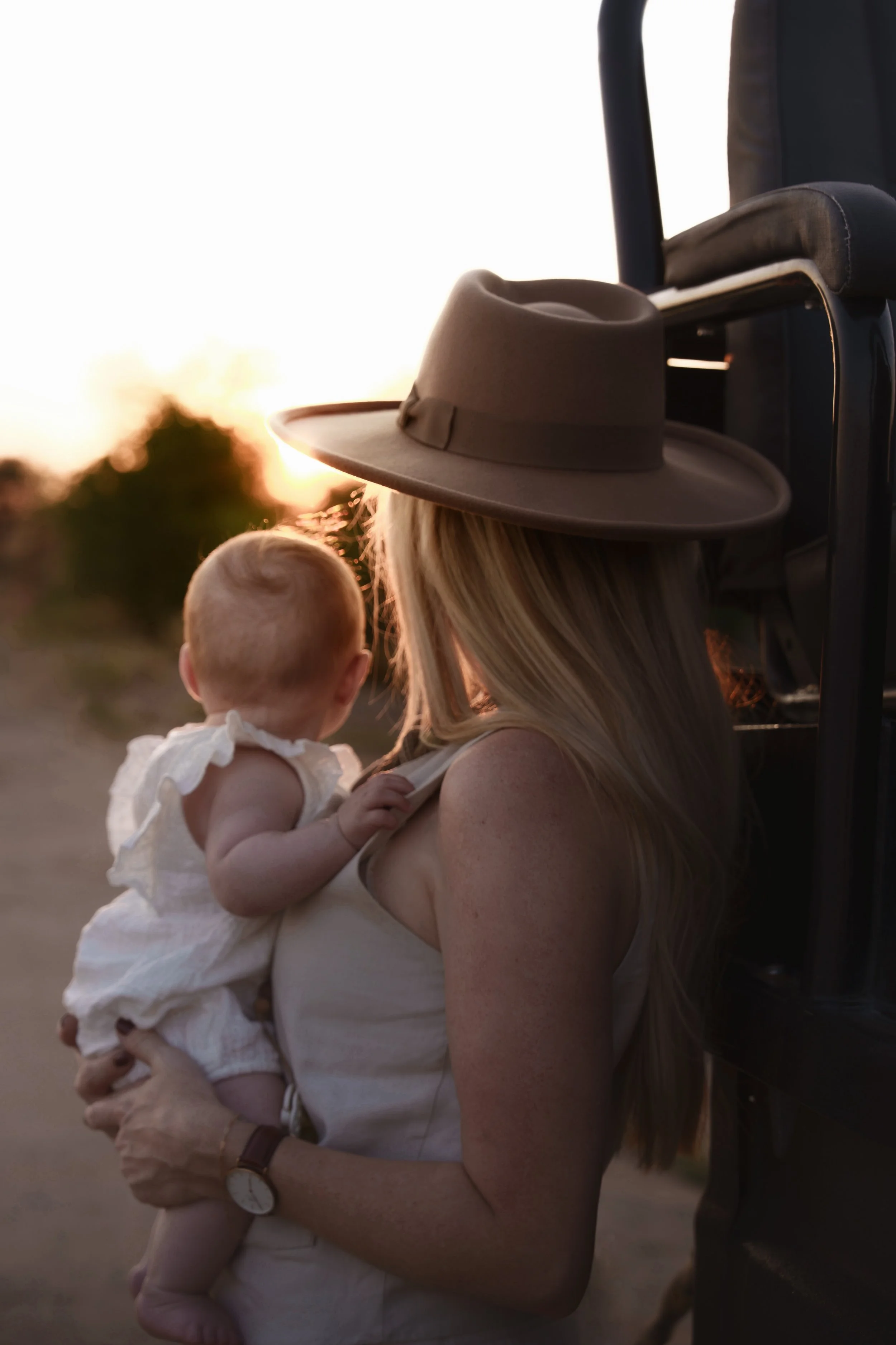 A woman with long blonde hair holding a young child, both facing away, during sunset outdoors; the woman has a beige hat on her lap and a black bracelet, while a person with a dark jacket is partially visible on the right.
