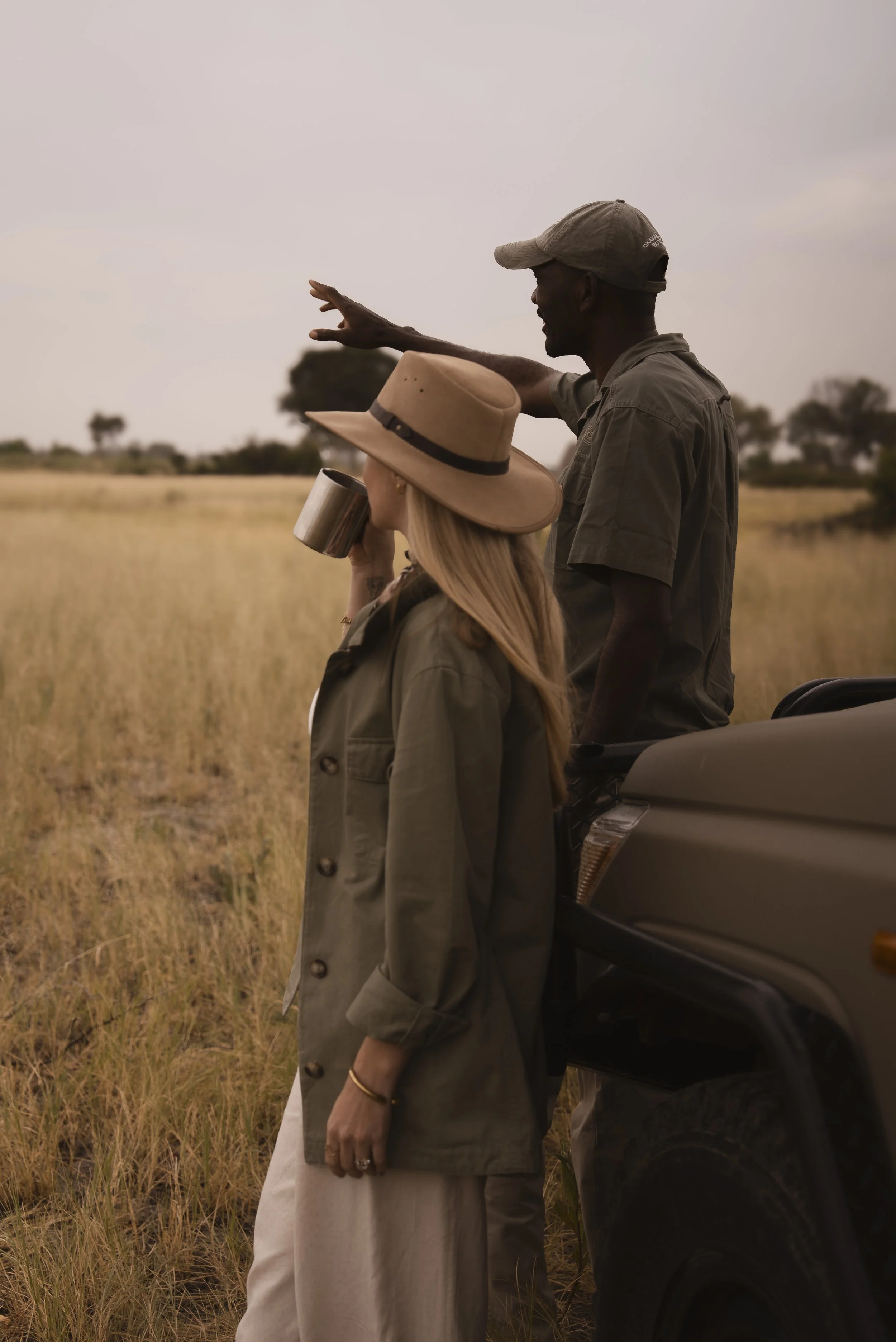 A man and a woman standing in a grassy field, with the man pointing into the distance while the woman holds a metal tumbler to her mouth, both wearing safari-style clothing and hats.