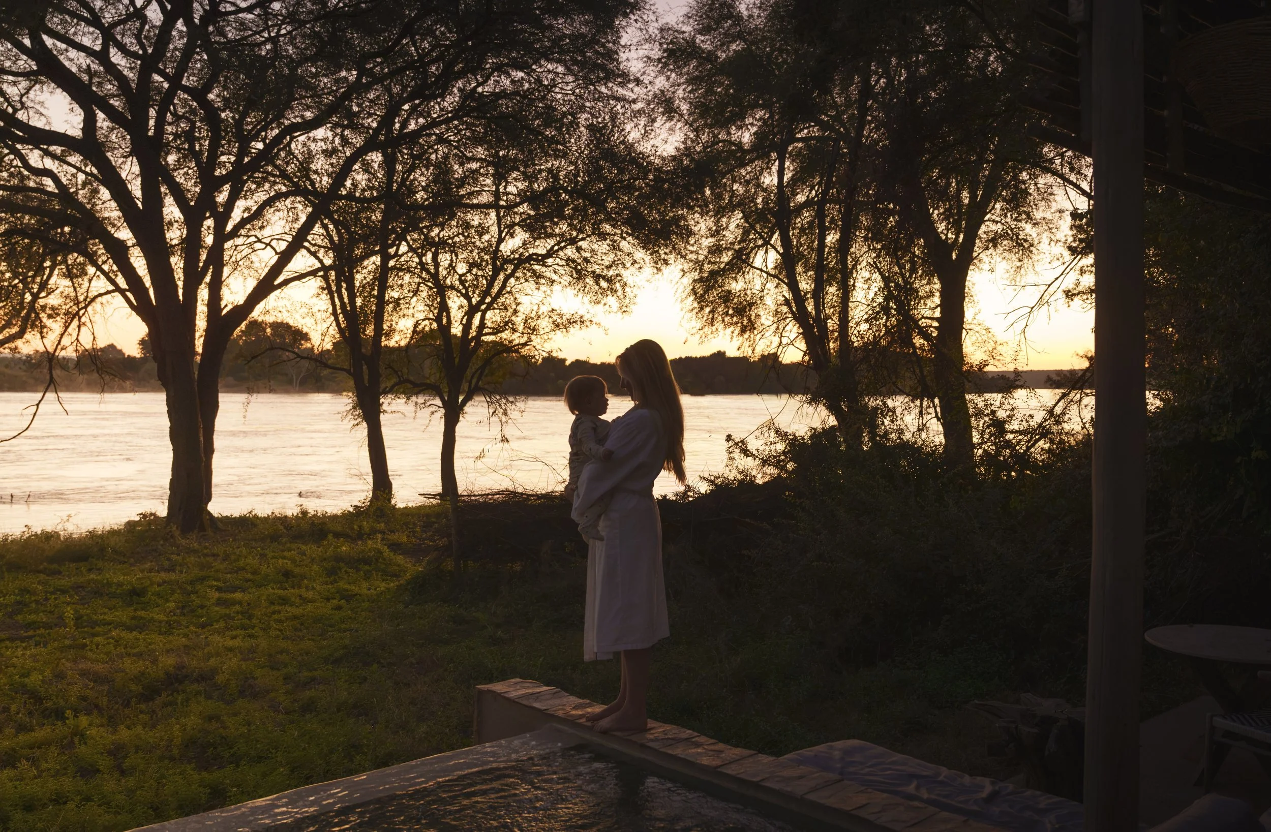 Mother holding young child in silhouette at sunset beside a river at a safari lodge