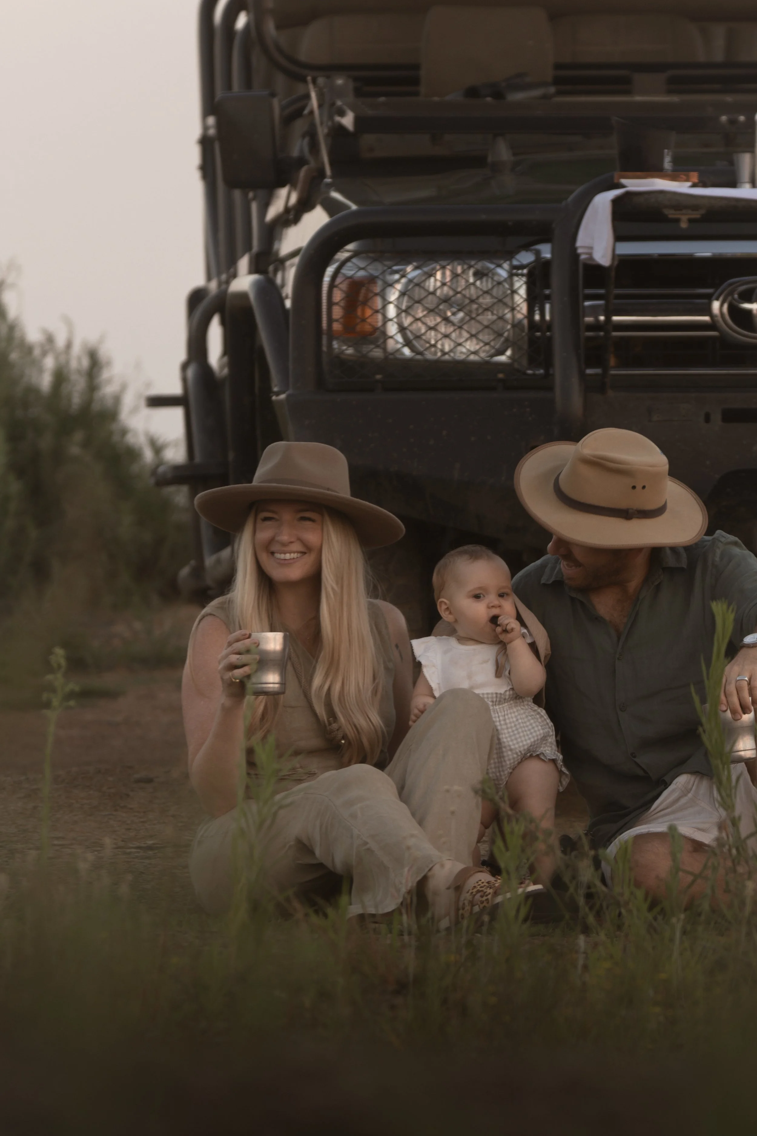 Parents sitting in the grass with their baby beside a safari vehicle at a sundowner stop