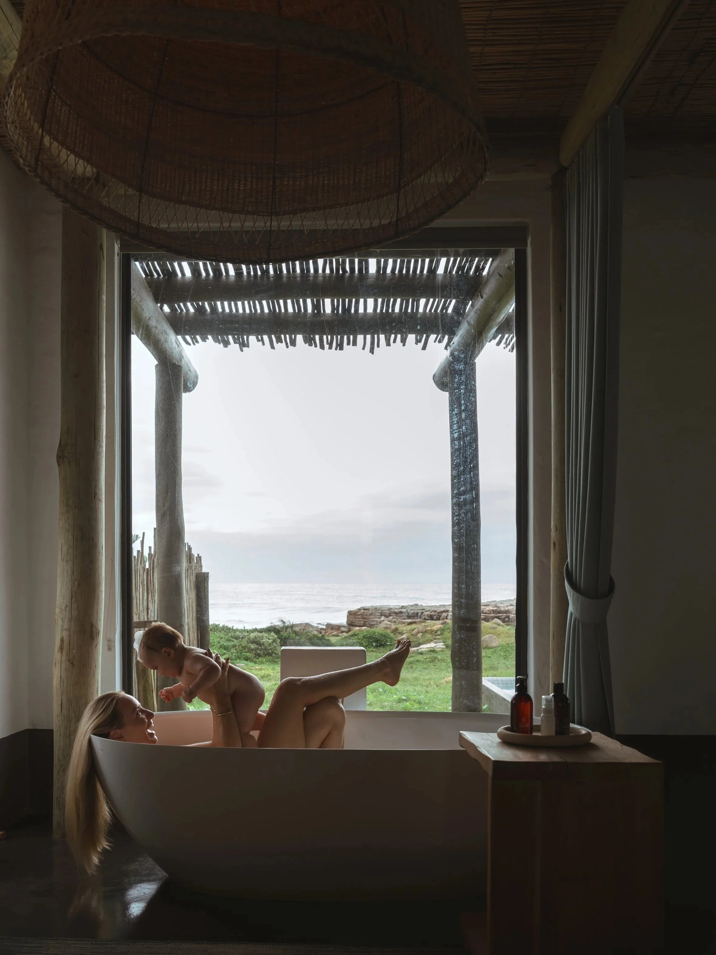 A woman bathing in a bathtub with a child on her legs, in front of a large window with a view of a beach and ocean outside, with a thatched roof visible through the window.