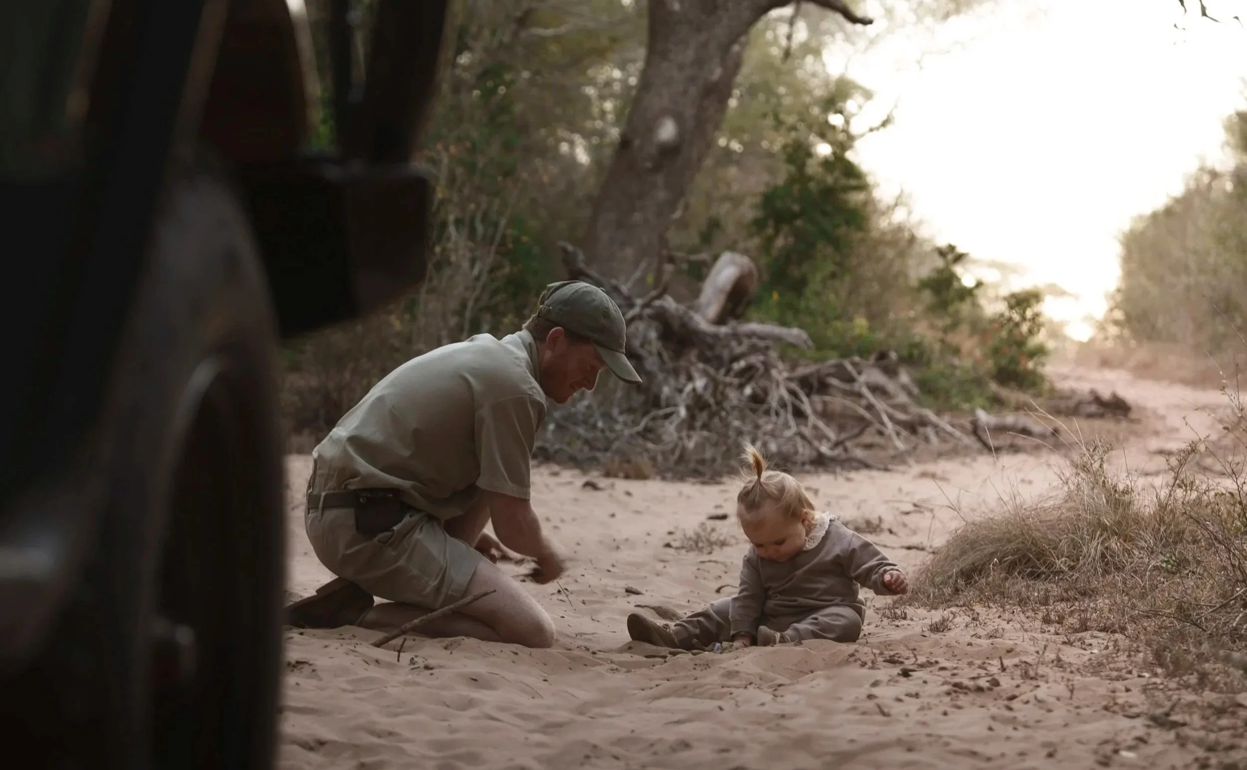 Safari guide kneeling down to play in the sand with a young child beside a game vehicle