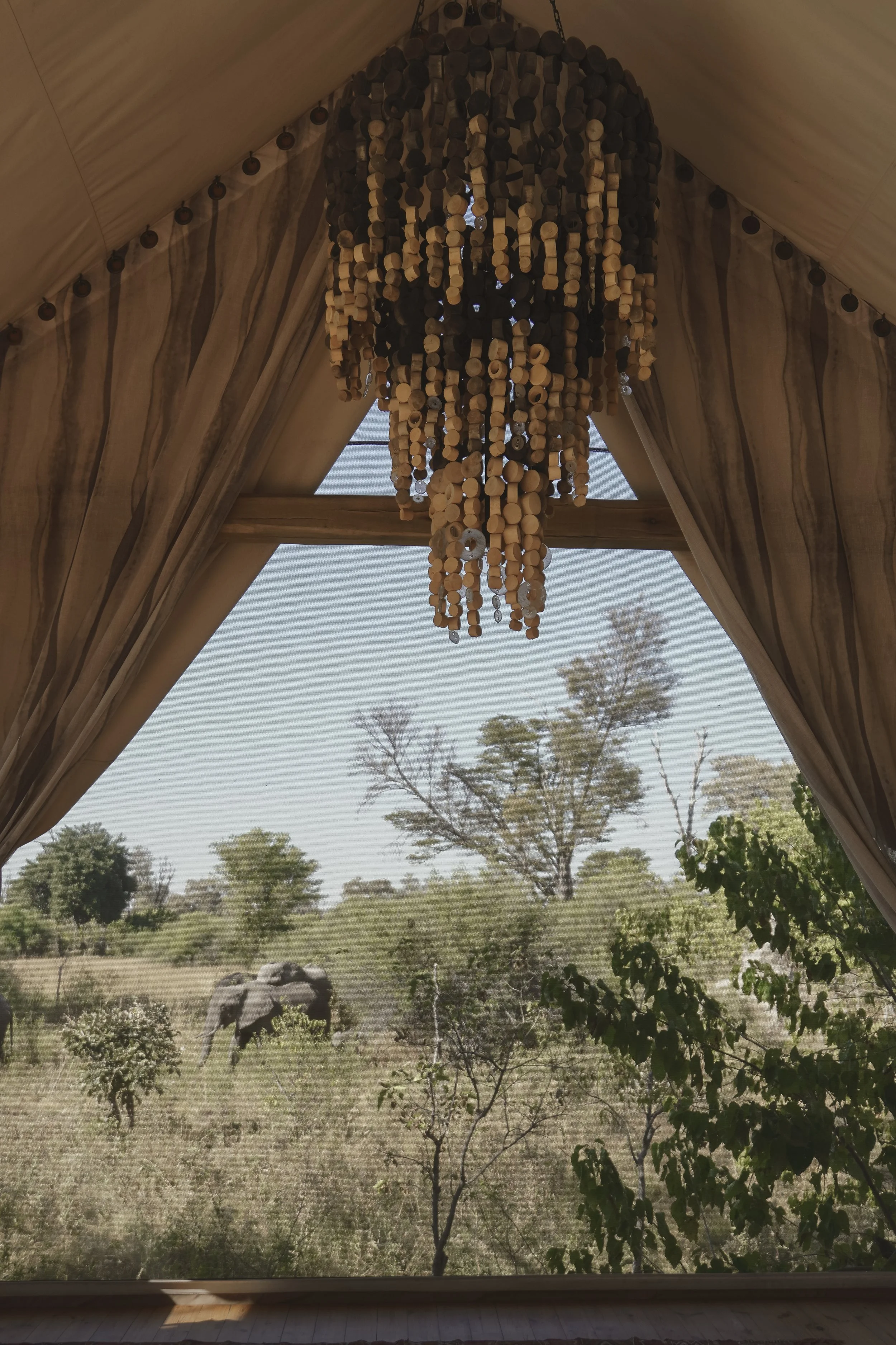 View through a window with beige curtains showing a landscape of trees, bushes, rocks, and a clear sky.
