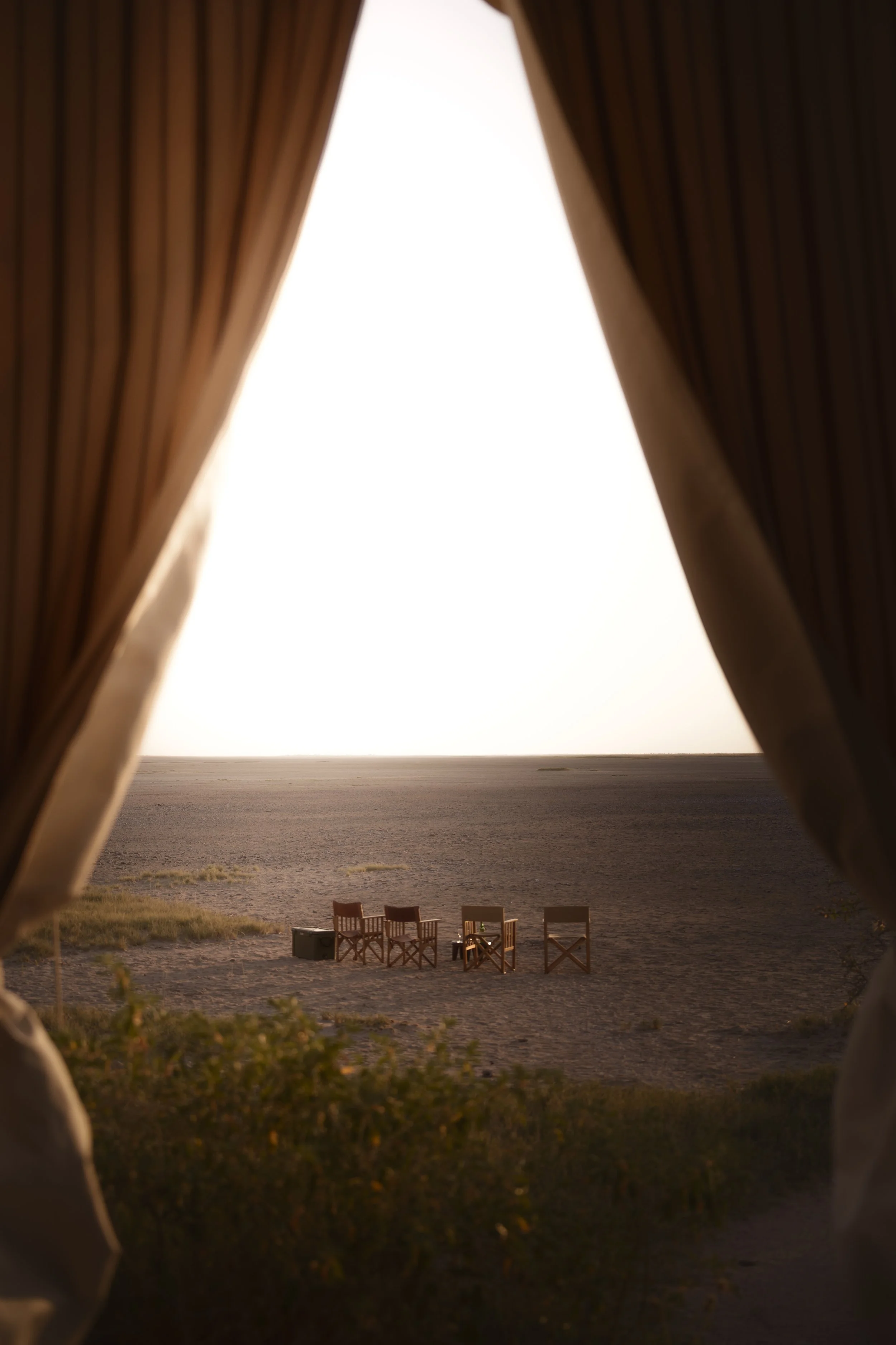 View through a slit or opening in a beige and brown tent showing chairs and a large open field or beach at sunset or sunrise.