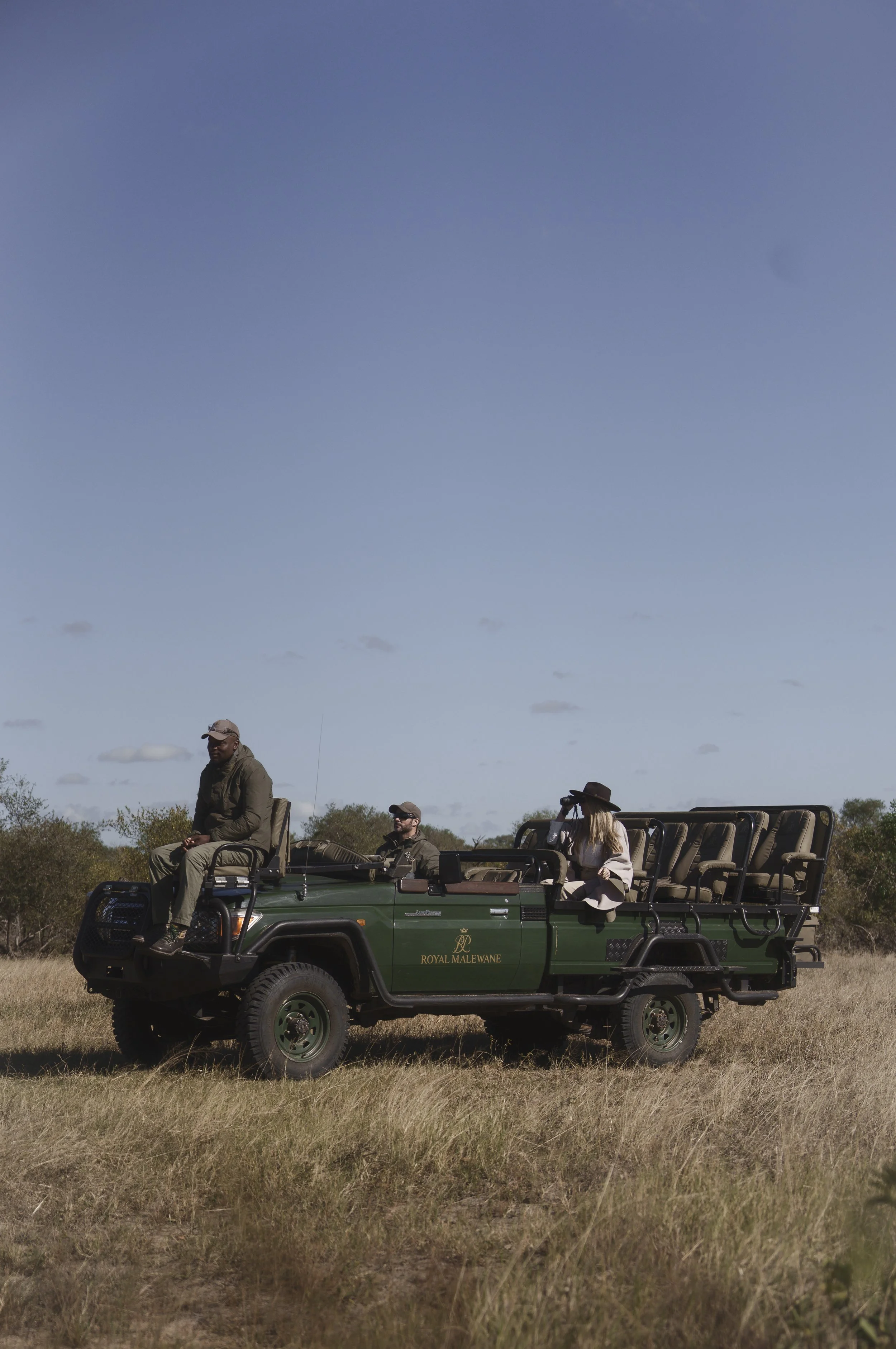 A group of people on a safari tour riding in an open green Royal Malewane vehicle across grassland under a blue sky with scattered clouds, some people wearing hats.