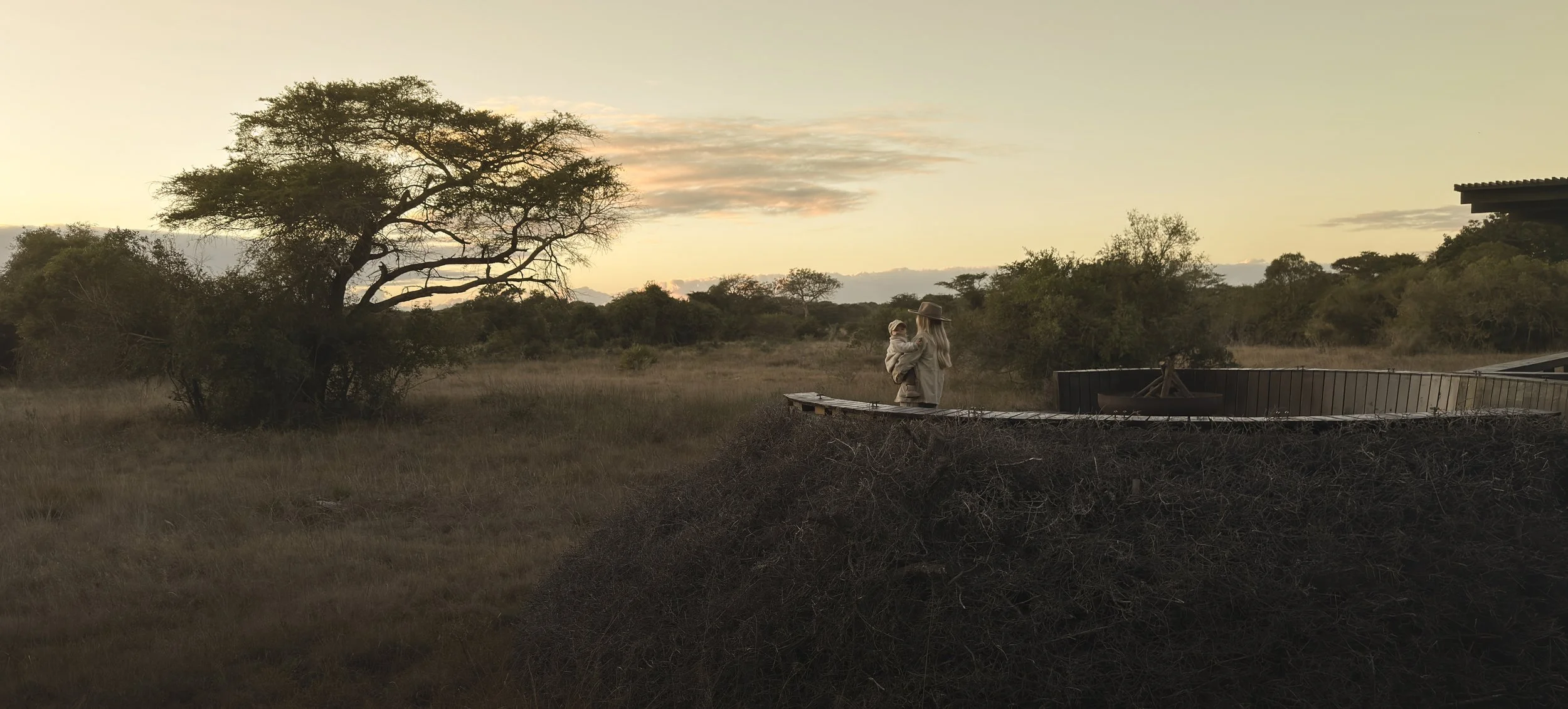 Mother holding a baby on a raised walkway at a safari camp at sunset