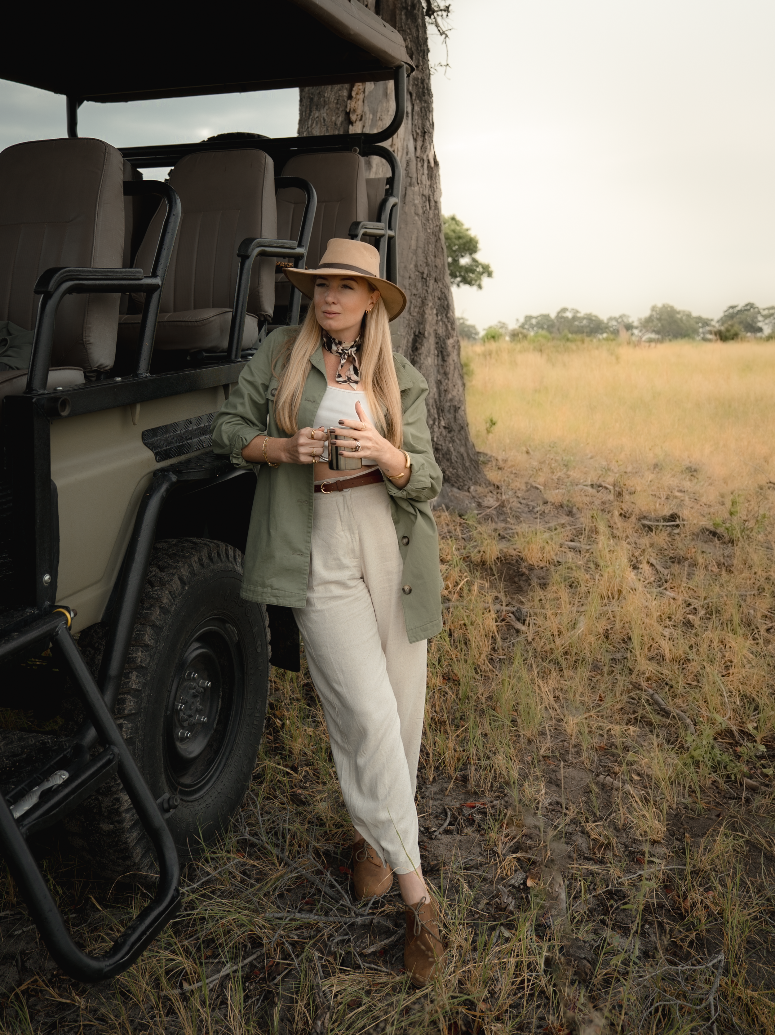A woman with long blonde hair, wearing a wide-brimmed hat, an olive green jacket, cream-colored pants, and brown boots, standing next to a safari vehicle in a grassy, open landscape with trees, holding a tumbler.