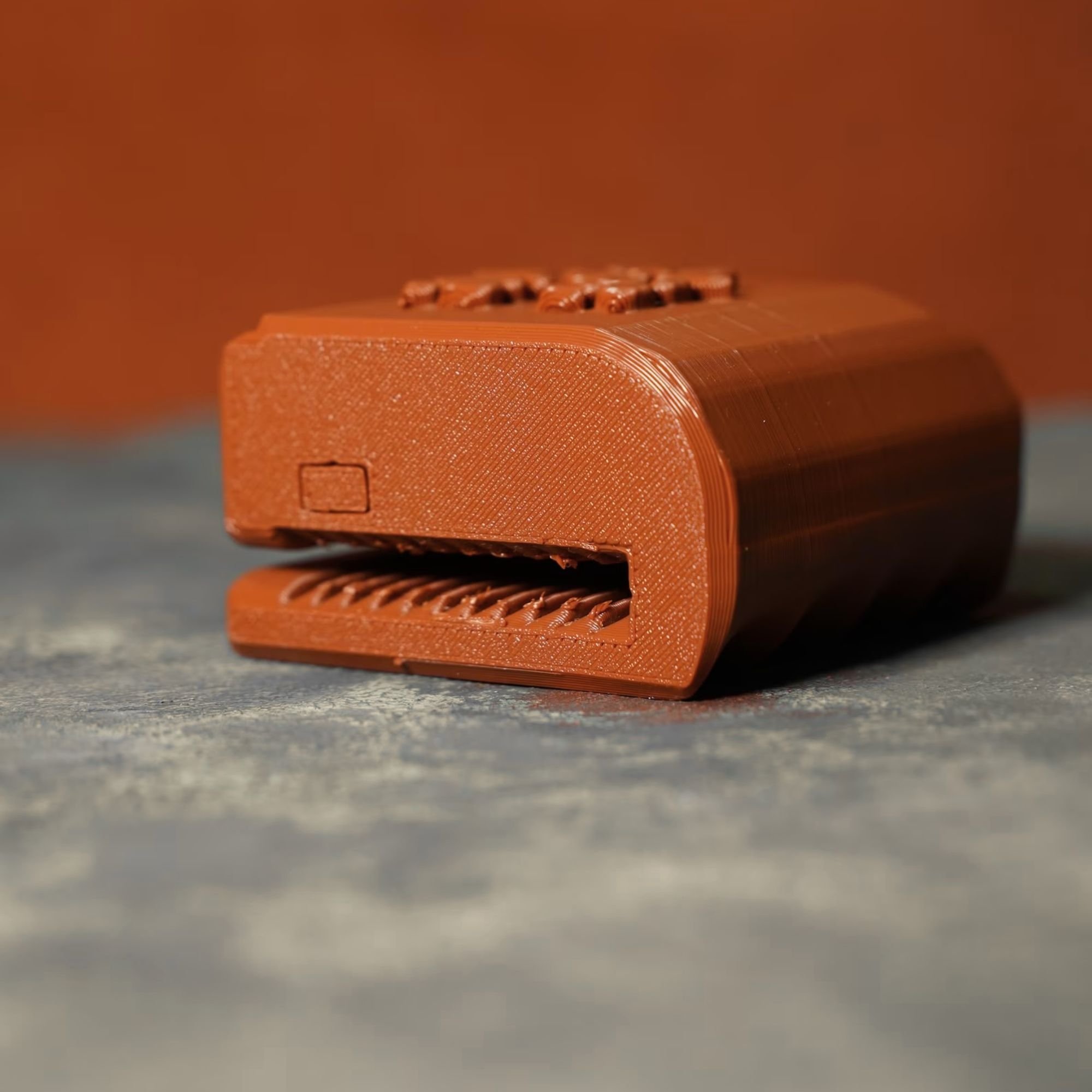 Close-up of a small orange plastic object with a textured surface, featuring ridges and rectangular indentations, resting on a grey surface with a plain background.