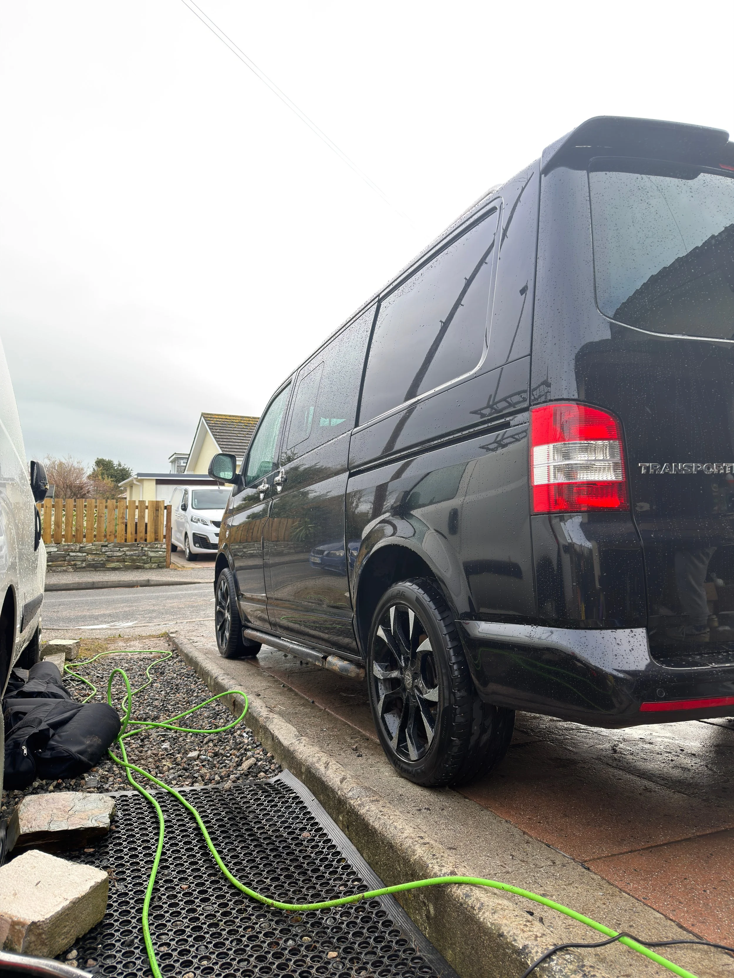 A black van parked on a driveway on a rainy day with water droplets on the surface. There are green extension cords and some paving stones near the curb.