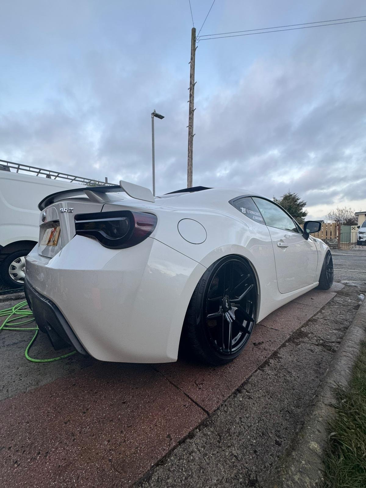White sports car parked on the side of the street with black wheels and a rear spoiler, under cloudy sky.