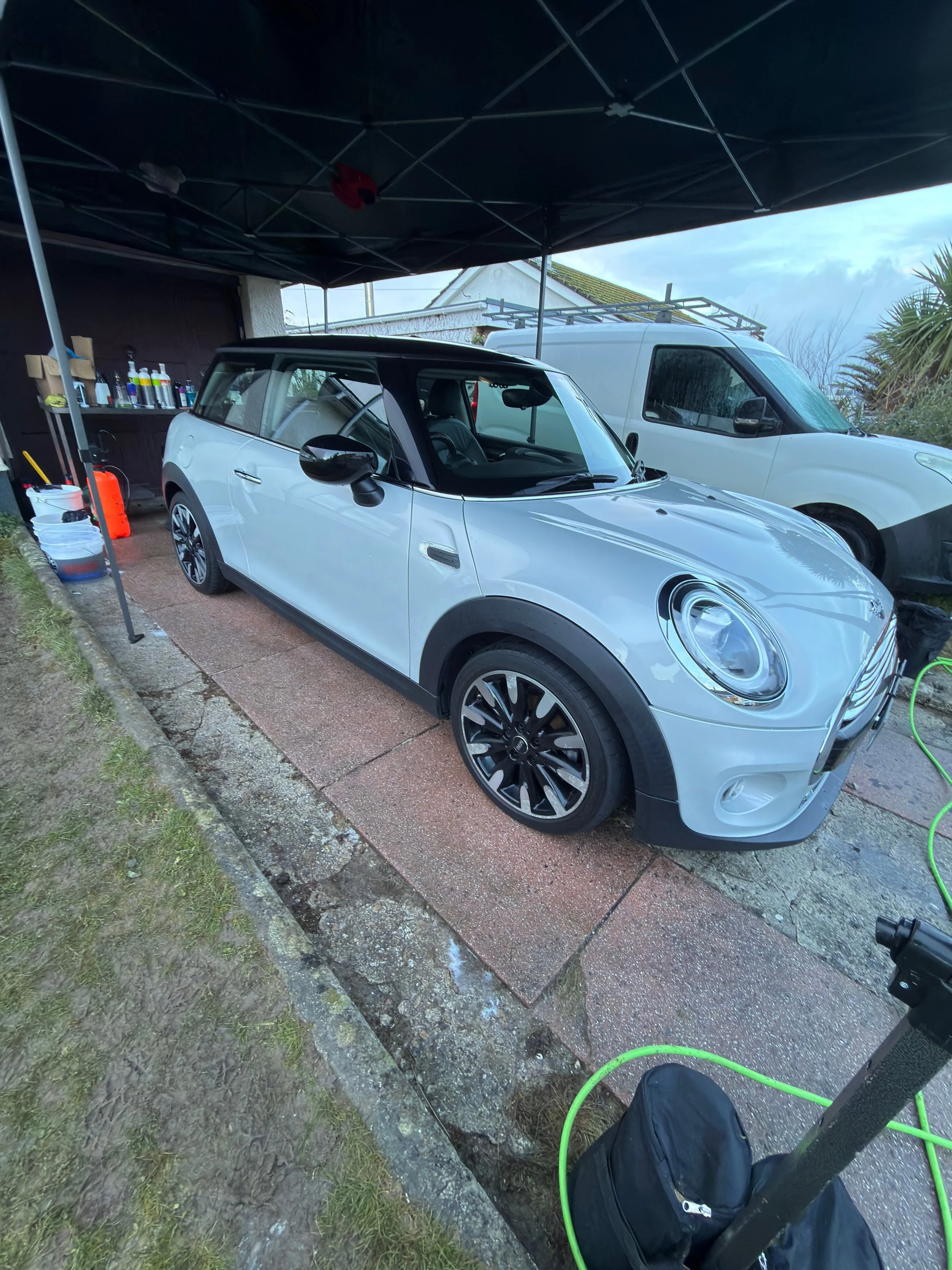 A white Mini Cooper car being washed or cleaned under a canopy, with a workbench and cleaning supplies on the left side, a white van parked behind it, and some greenery in the background.