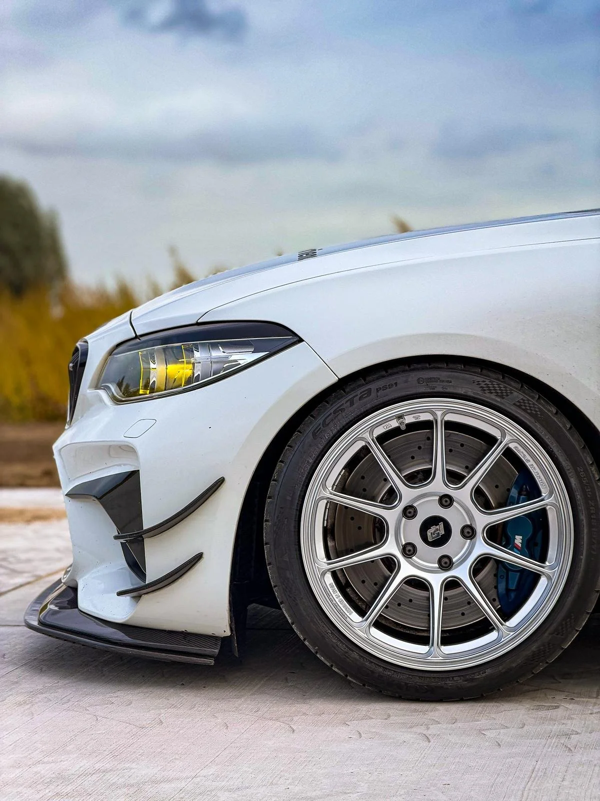 Close-up of the front part of a white sports car, showing a detailed wheel, brake caliper, and headlight, parked outdoors.
