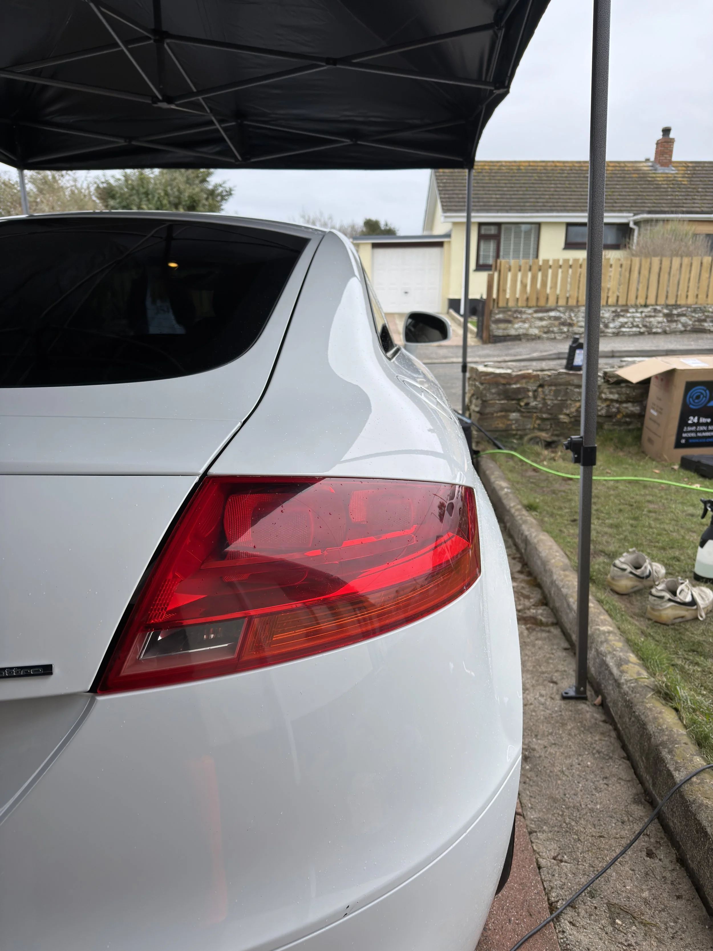 The rear side of a silver car parked under a canopy, with a red tail light visible, in a residential driveway with shoes and a box nearby.