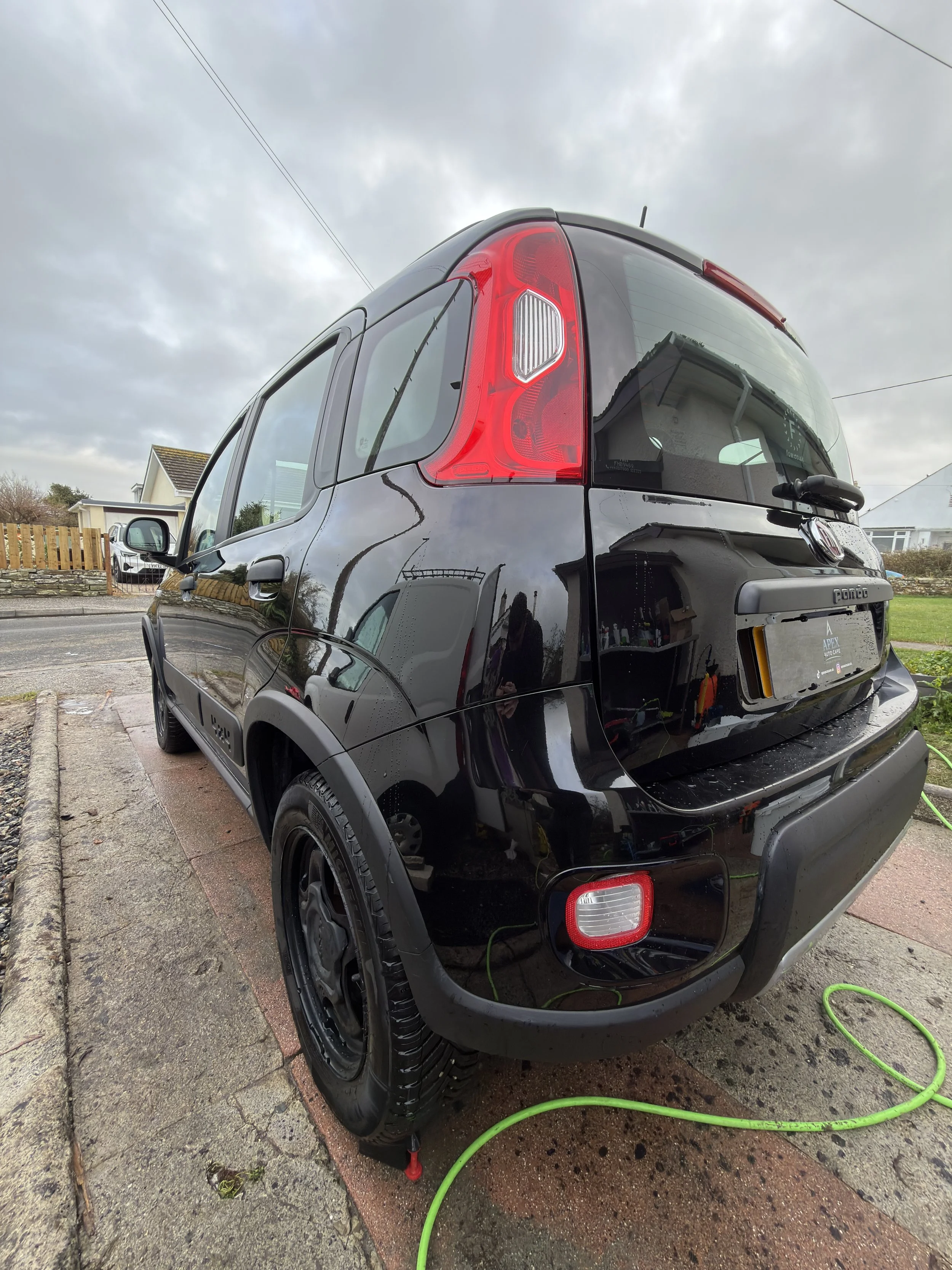 Black Fiat Panda parked on driveway on a cloudy day, with green hose on the ground.