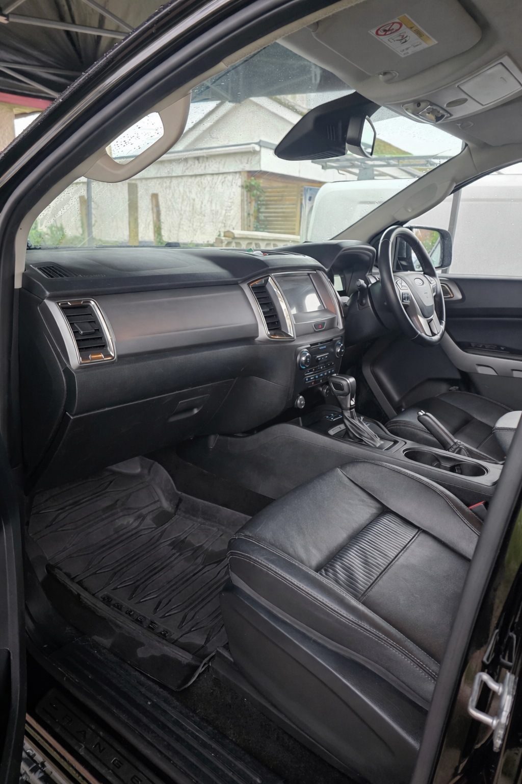Interior of a black pickup truck with leather seats, dashboard, steering wheel, gear shift, and front passenger seat, parked outdoors during rainy weather.