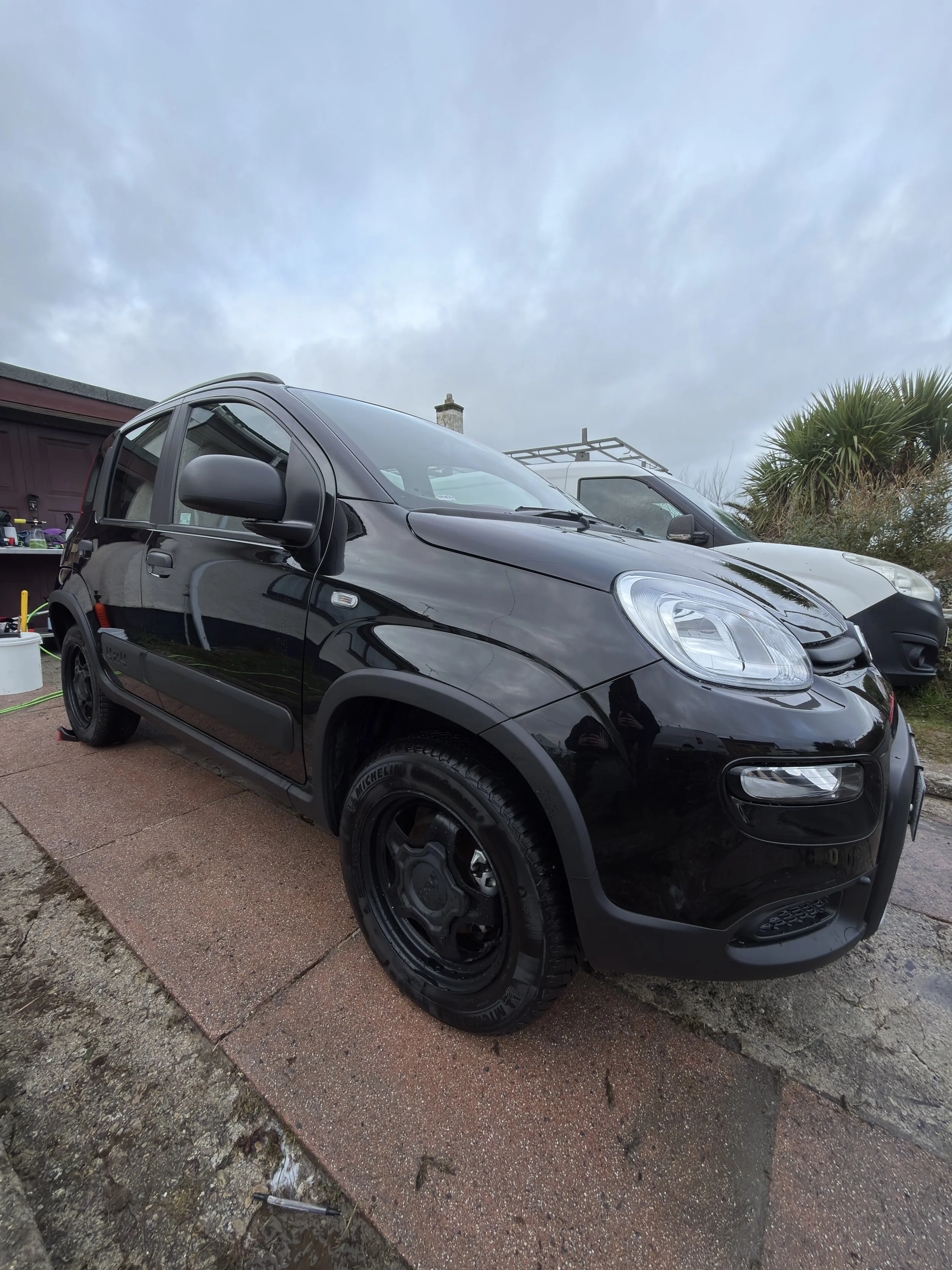 Black compact car parked on a driveway next to a white vehicle, with a cloudy sky and some greenery in the background.