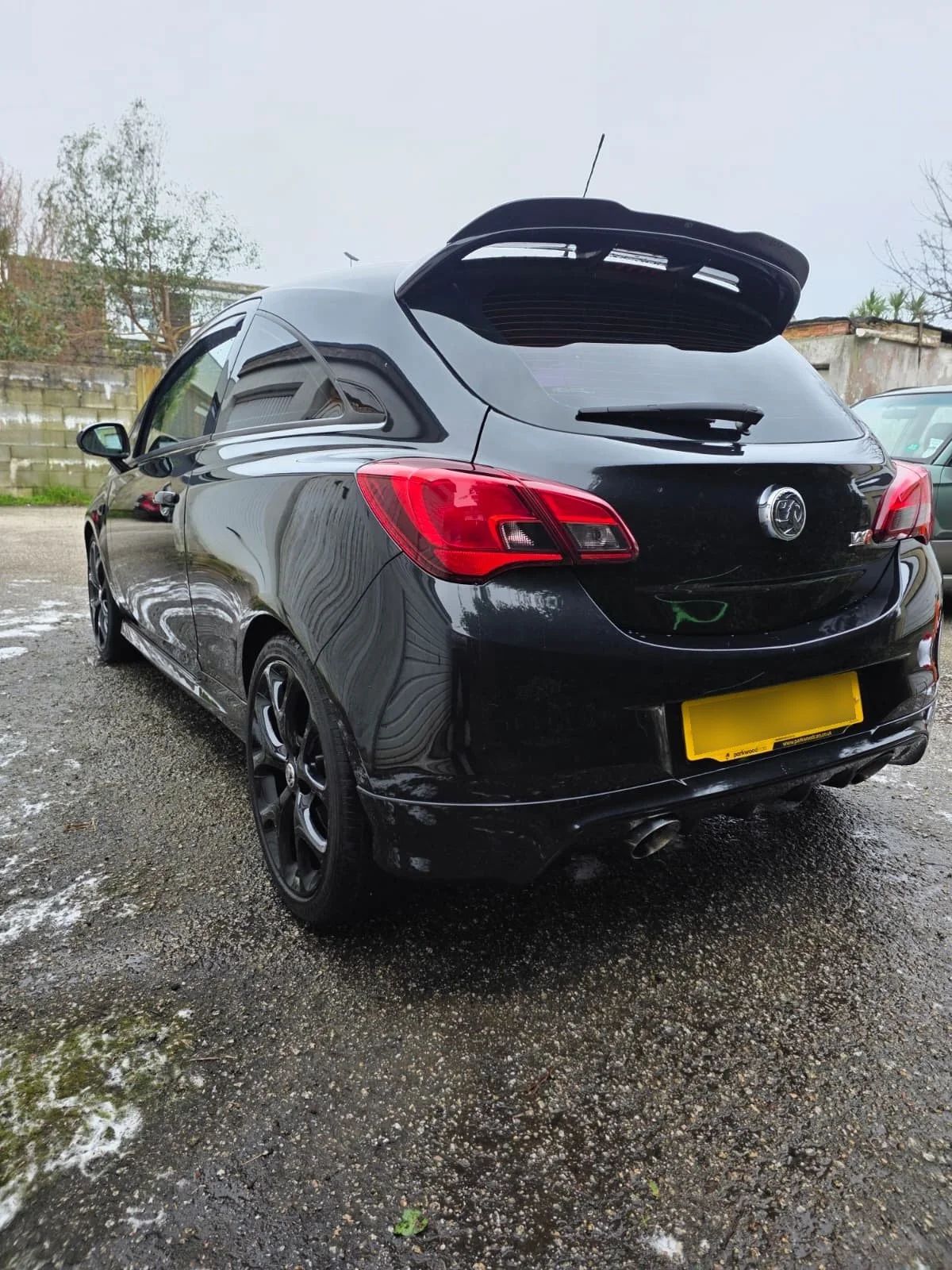 Black hatchback car parked on wet gravel, with a yellow license plate, dark tinted windows, and a rear spoiler, outdoors during daytime.