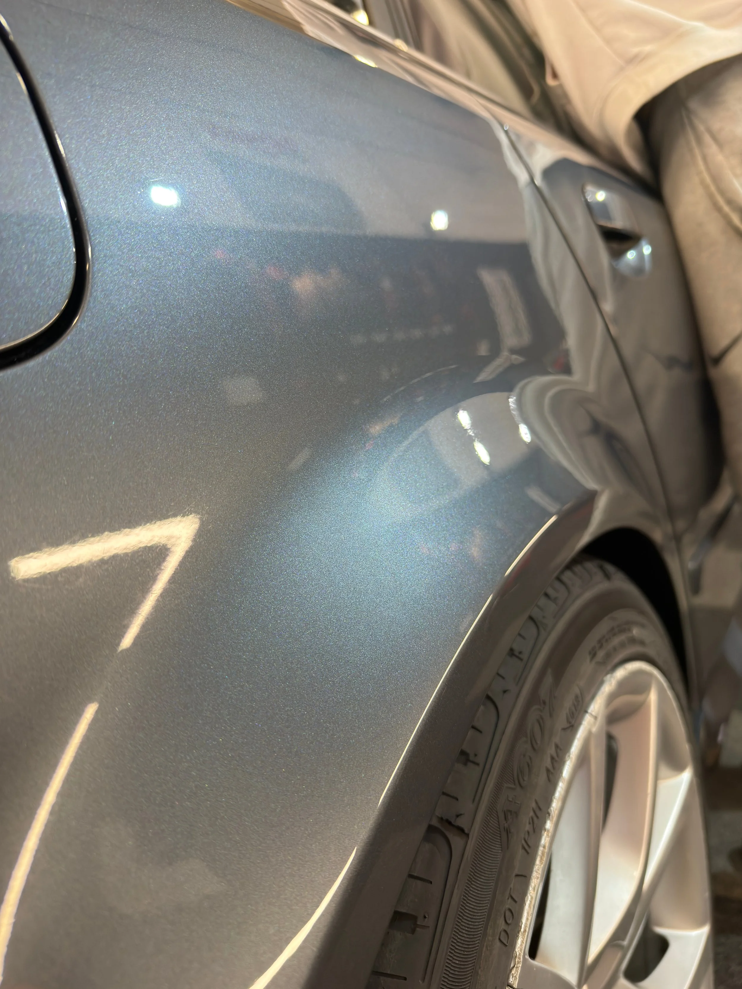 Close-up of a metallic gray car's body, showing a shiny surface with reflections of lights and surroundings.