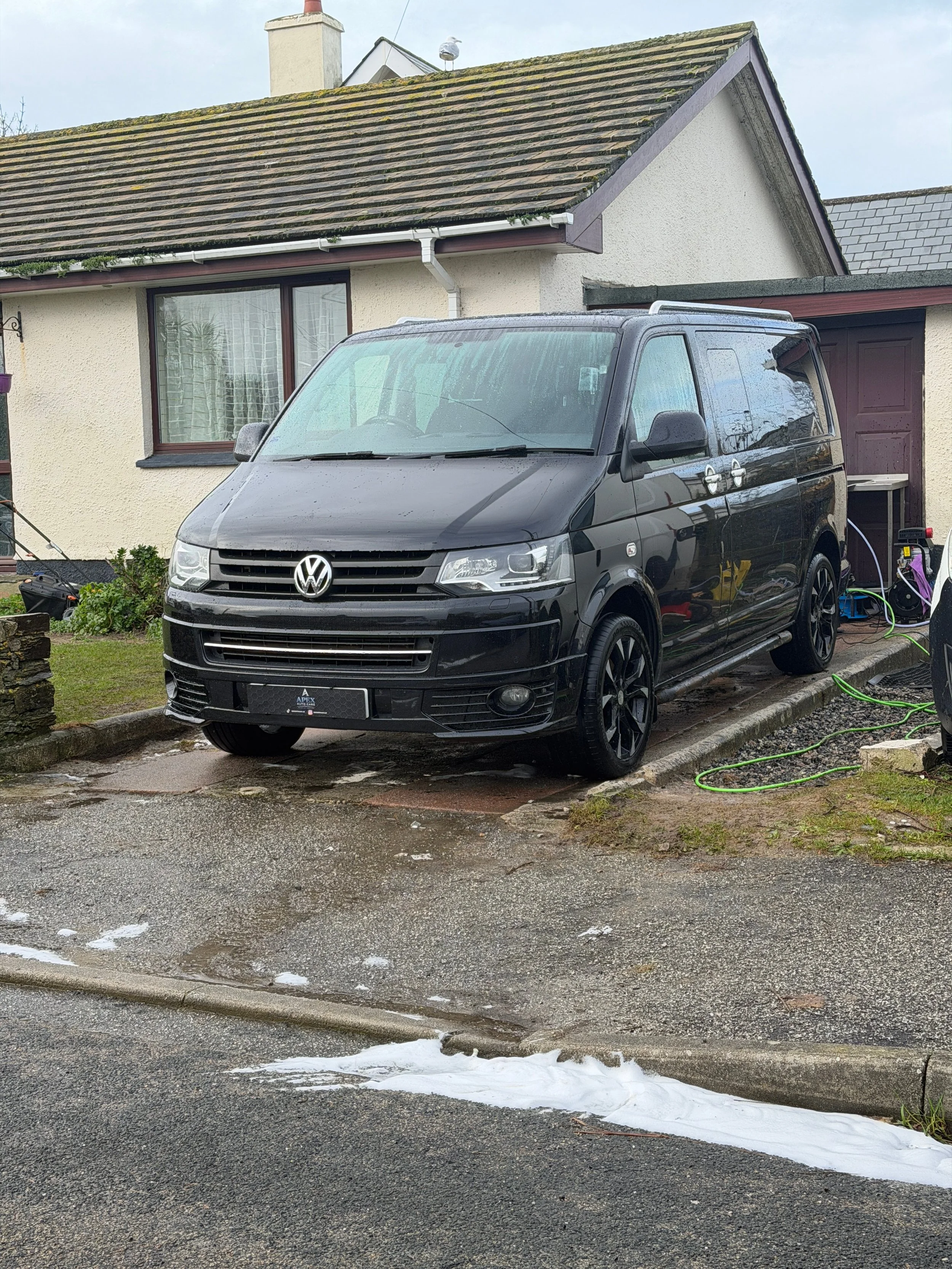 A black Volkswagen van parked on a driveway in front of a house, with soap and water on the vehicle and on the ground, indicating it is being washed.