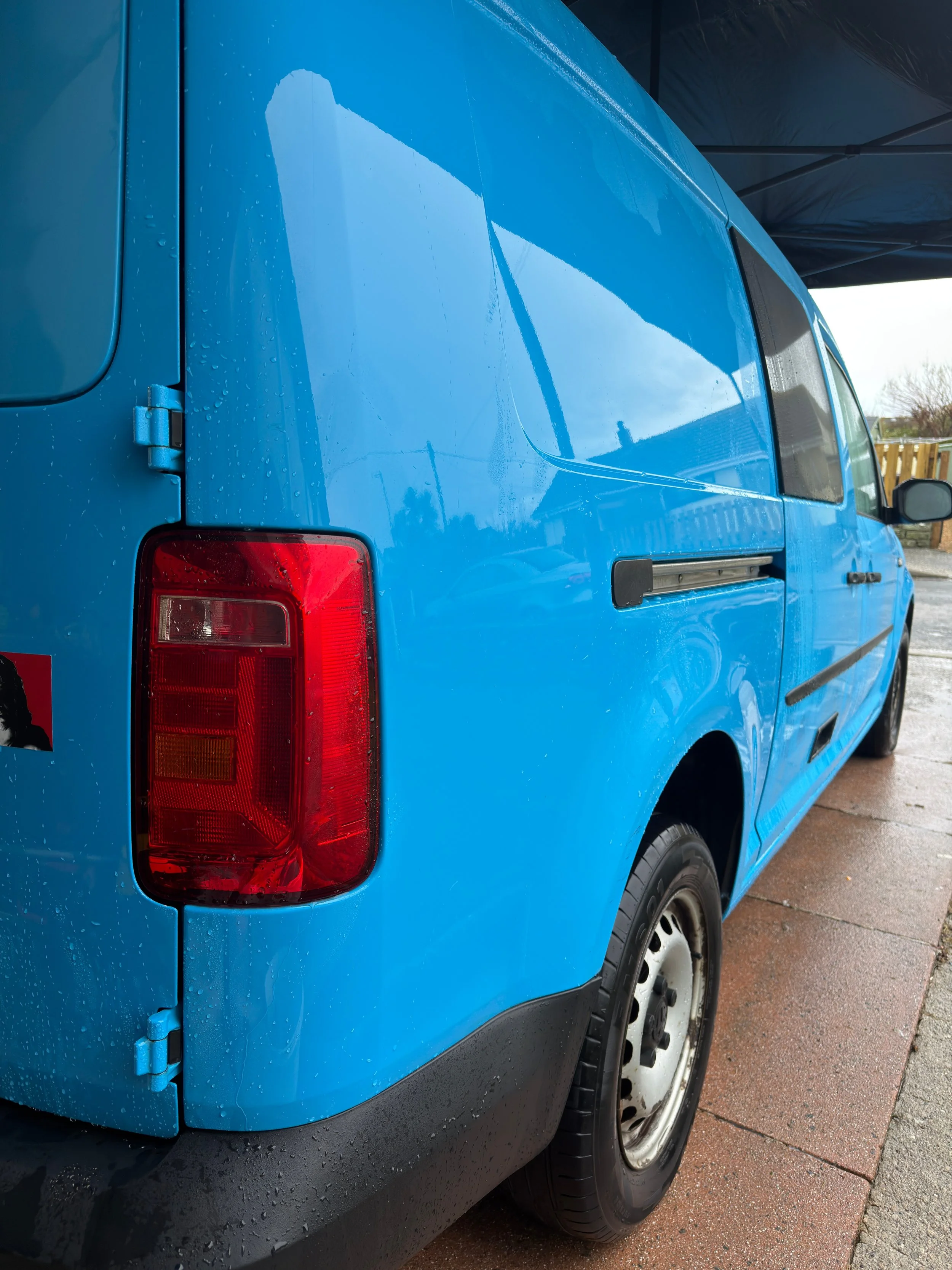 Blue van parked outdoors under a canopy with water droplets on its surface.