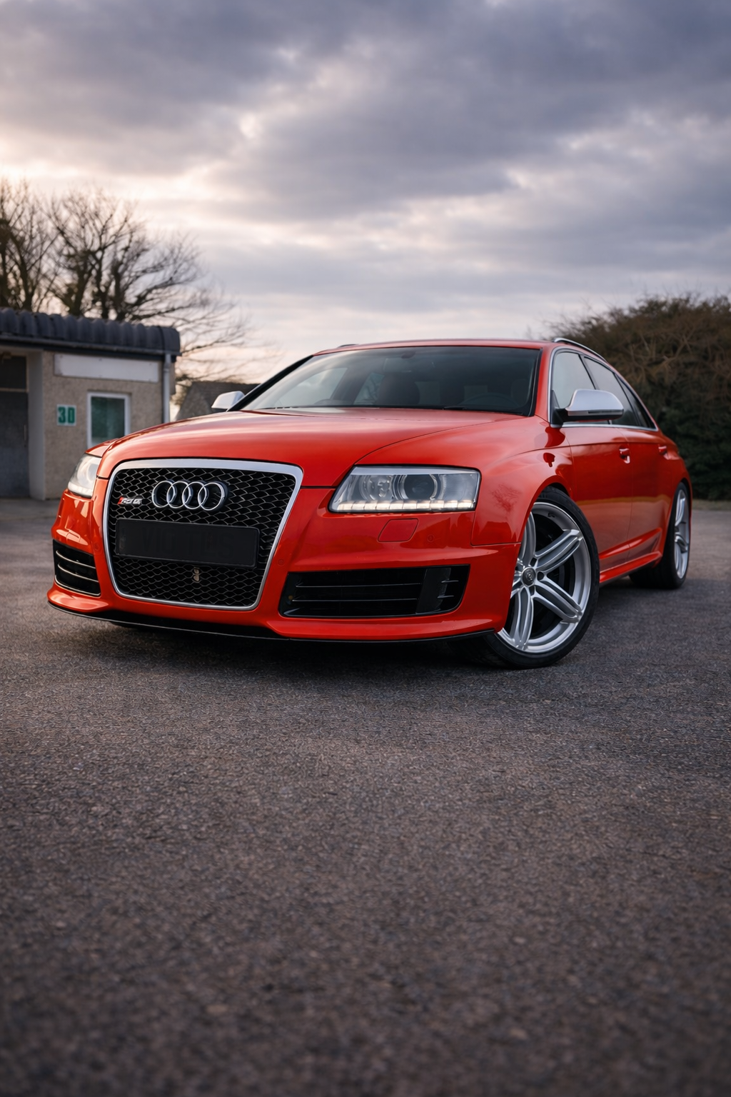 Red Audi sedan parked on asphalt with cloudy sky and trees in the background.