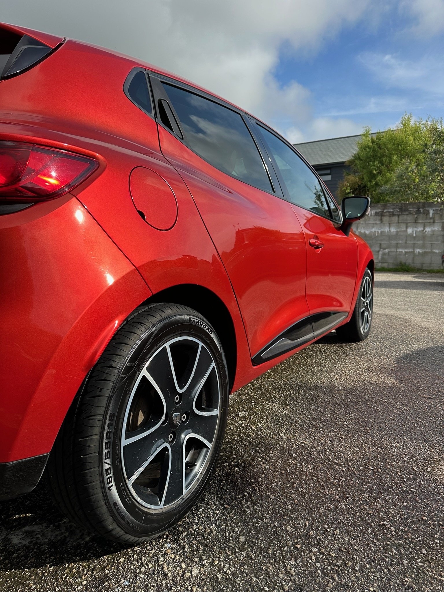 Red hatchback car parked on asphalt with a cloudy sky and trees in the background.