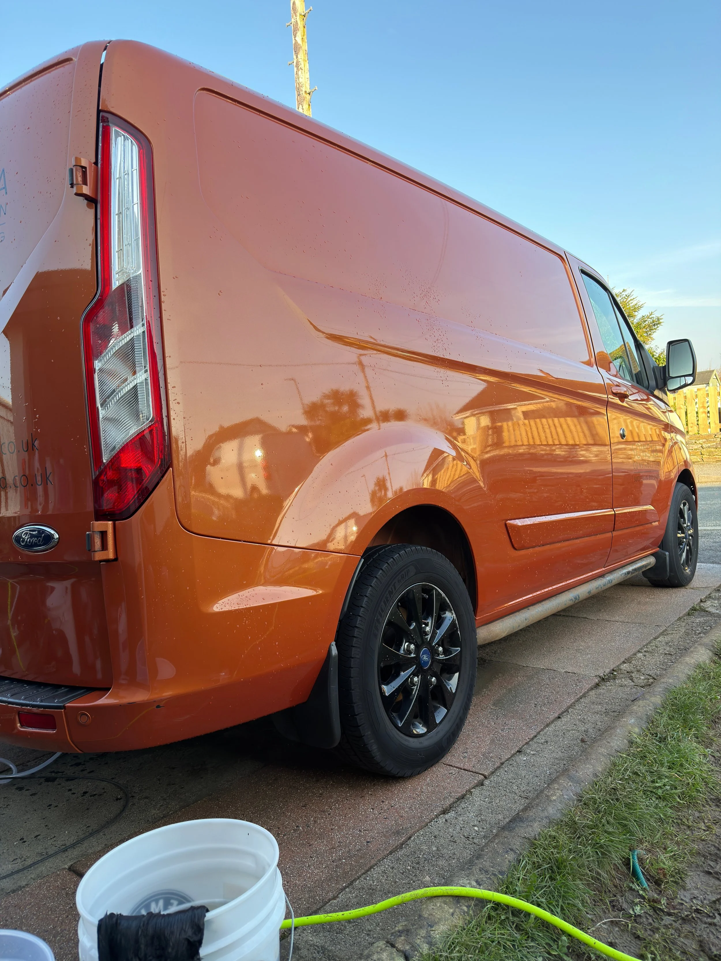 Orange panel van parked on a driveway with cleaning supplies nearby, reflecting the sky and surroundings in its shiny surface.
