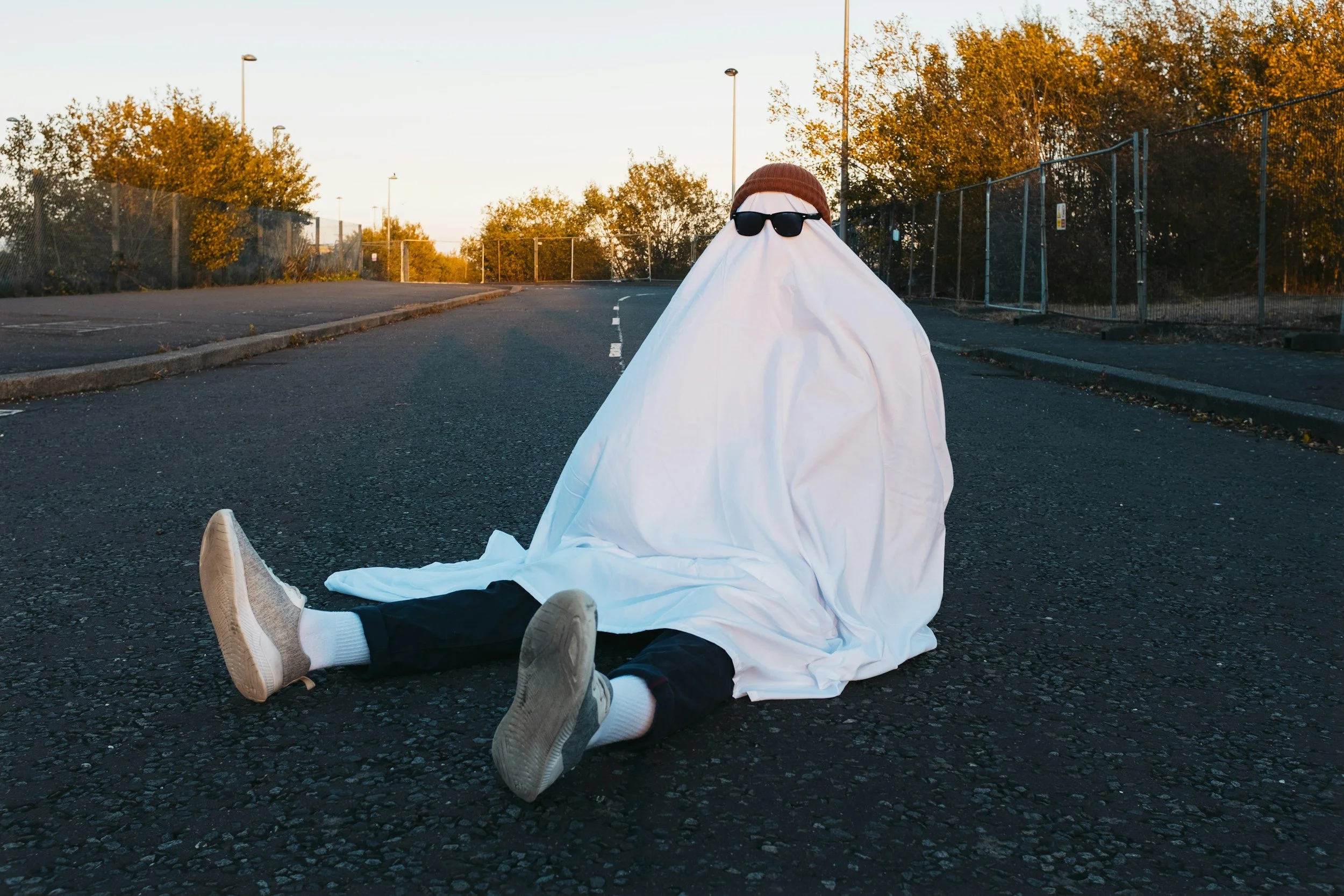 Person sitting on the street wearing a white sheet with sunglasses and a brown cap, with their legs and shoes visible, during sunset or sunrise.