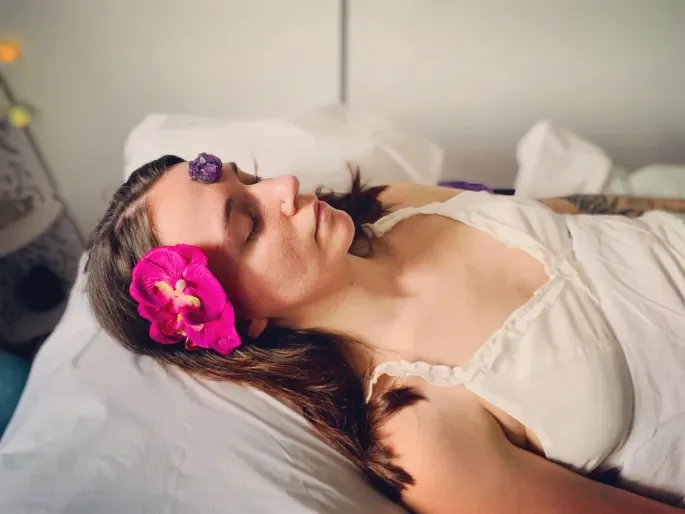 A woman lying in bed with her eyes closed, wearing a white tank top and floral hair accessories, including a pink orchid near her head.