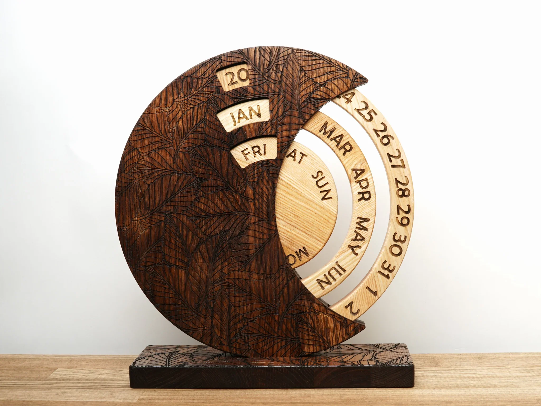 Wooden calendar with circular date, day, and month displays, and carved leaf pattern on dark and light wood sections.