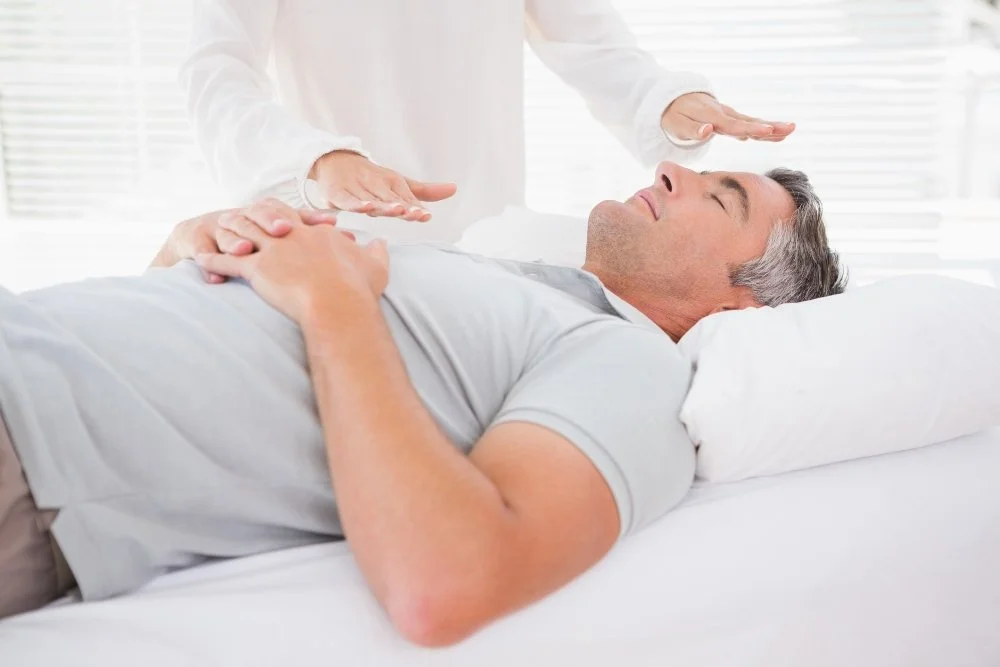 A man lying in bed with his eyes closed, receiving a holistic or Reiki healing session from a person whose hands are hovering above his chest.
