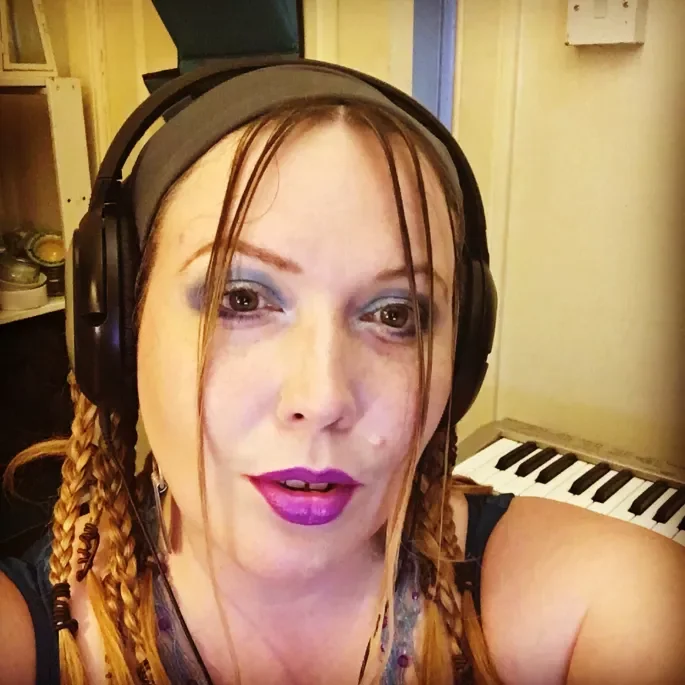 A woman with braided hair wearing large headphones, purple makeup, and a dark tank top. She is in a room with a keyboard in the background.