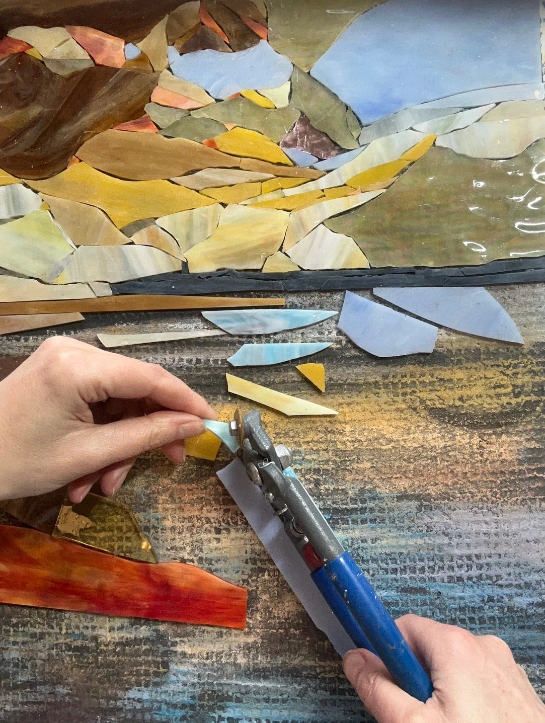 Person assembling colourful mosaic glass pieces on a wooden work surface, using a glass cutter.