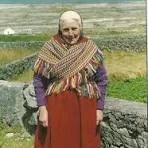 A historical photo of an elderly woman with a headscarf standing outdoors near a field.
