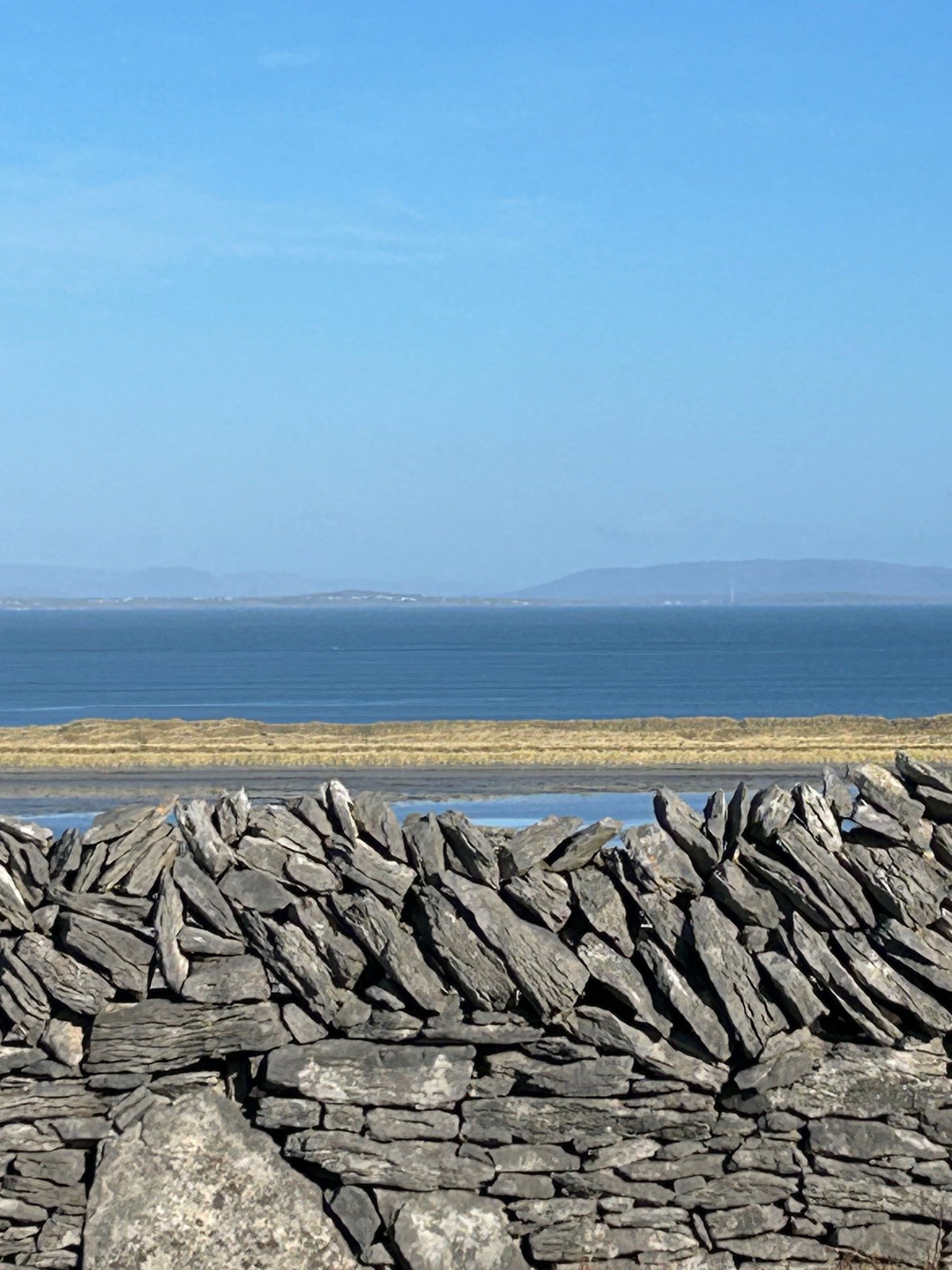 A stone wall with a view of a body of water and distant land under a clear blue sky.