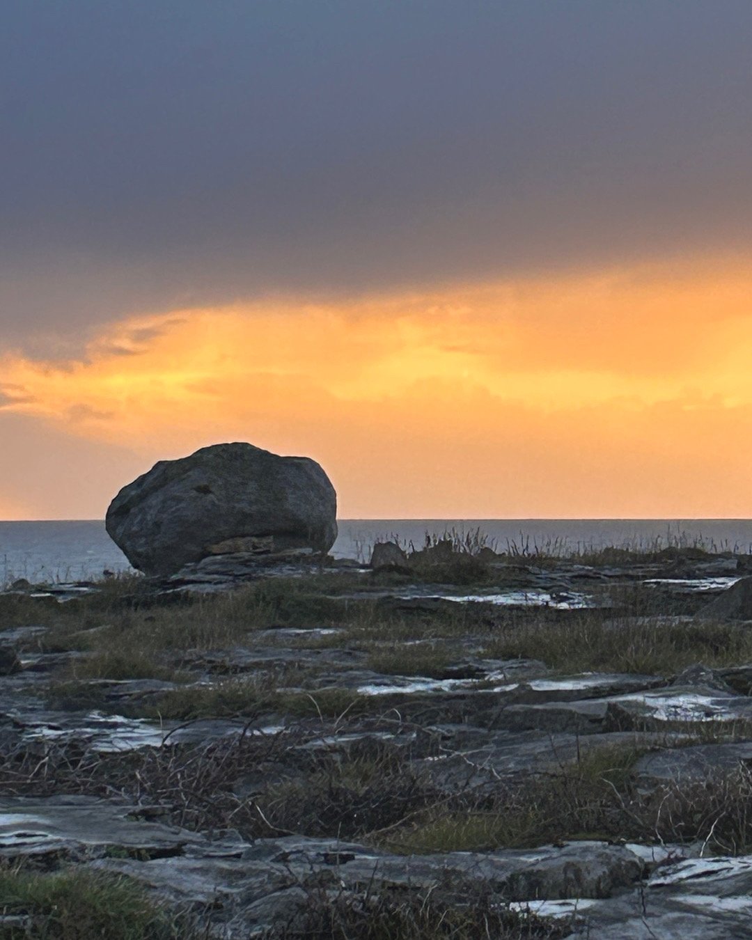 A large rock on a grassy and rocky shoreline at sunset with an orange and yellow sky.