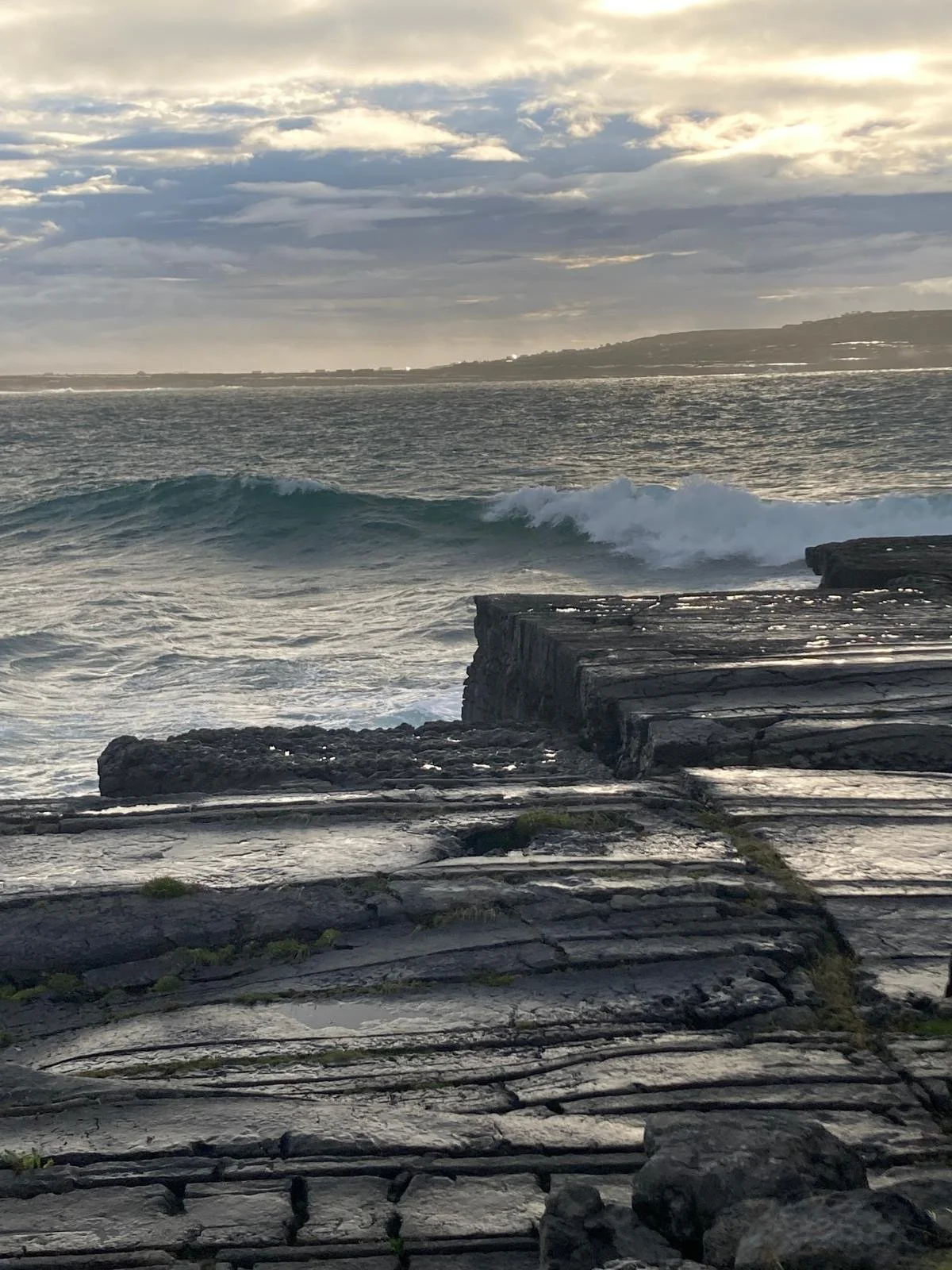 A rocky shoreline with a wave breaking in the ocean under a cloudy sky, with land in the background.