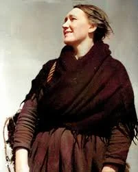A historical photo of a woman smiling while looking to her right, wearing a dark shawl and a long dress.