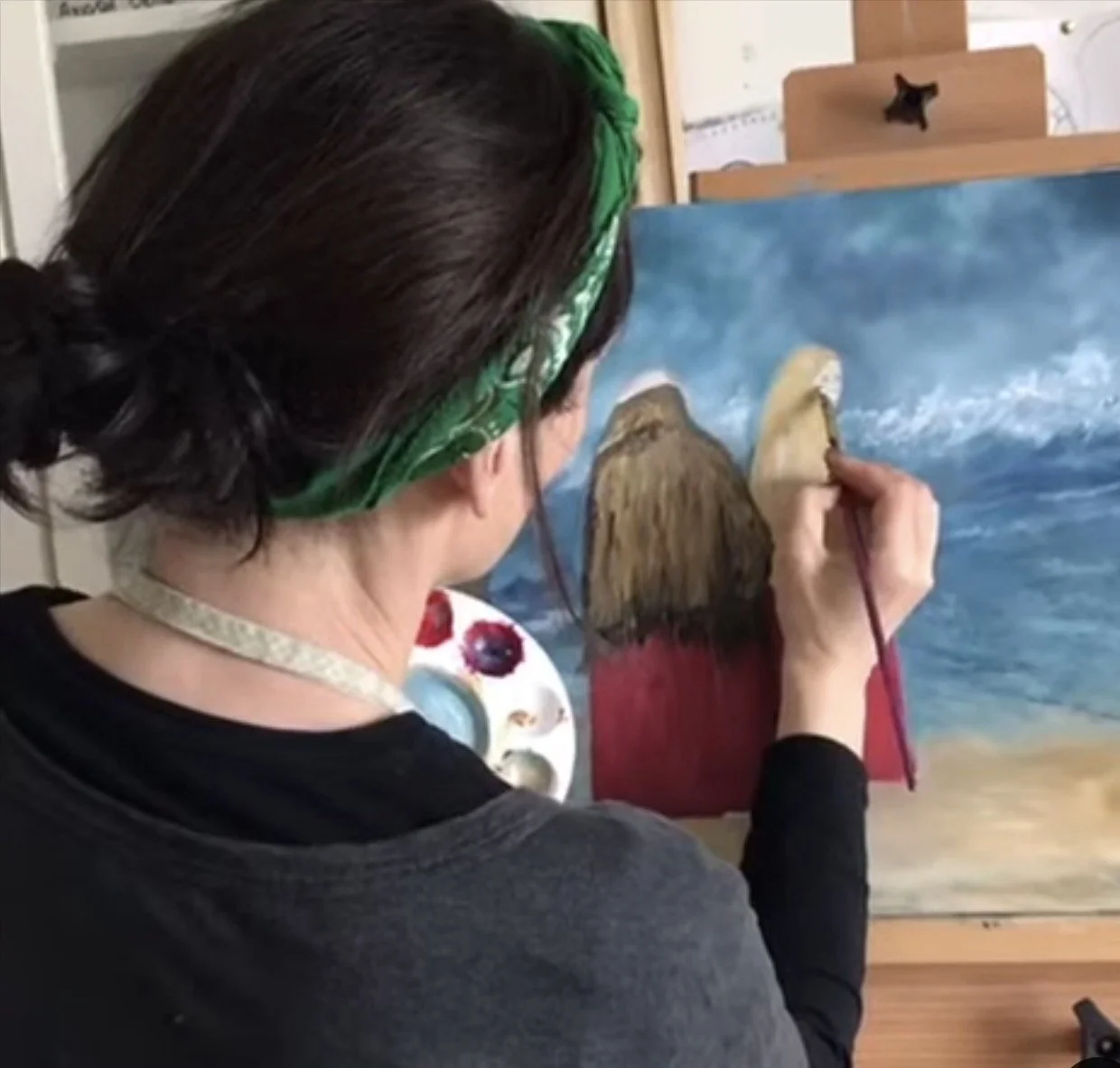 An artist is working on a painting of two women wearing shawls, with art supplies nearby.