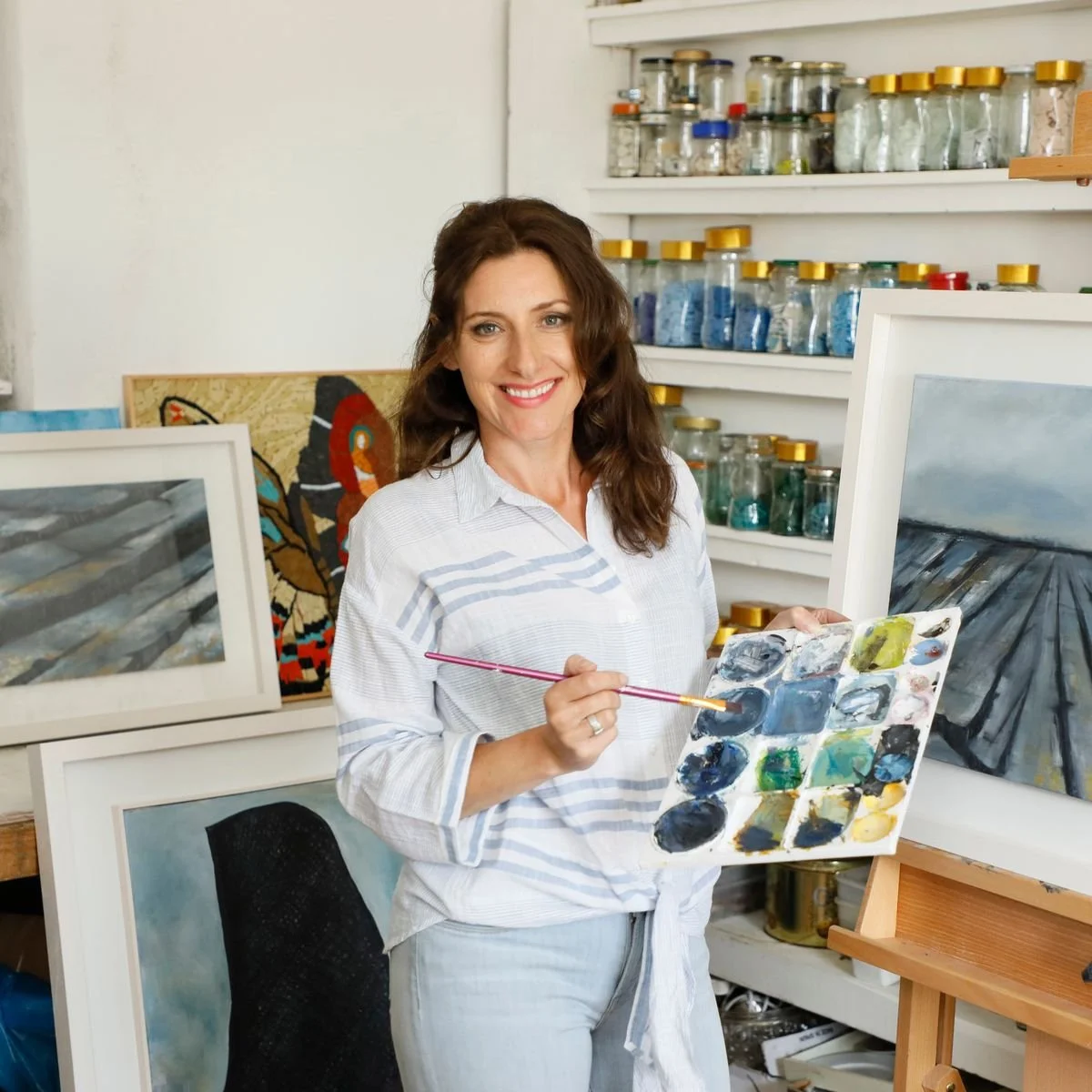 A woman standing in an art studio, holding a painter's palette with various colors and a paintbrush, smiling at the camera. Behind her are shelves with jars of paint and multiple framed paintings.