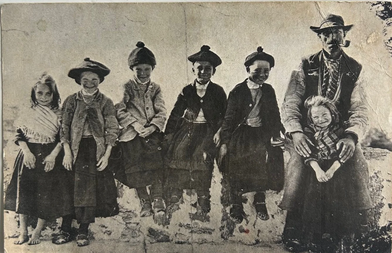Old black and white photograph of six children and one man, all wearing vintage clothing, sitting or standing outdoors on a rough stone surface.