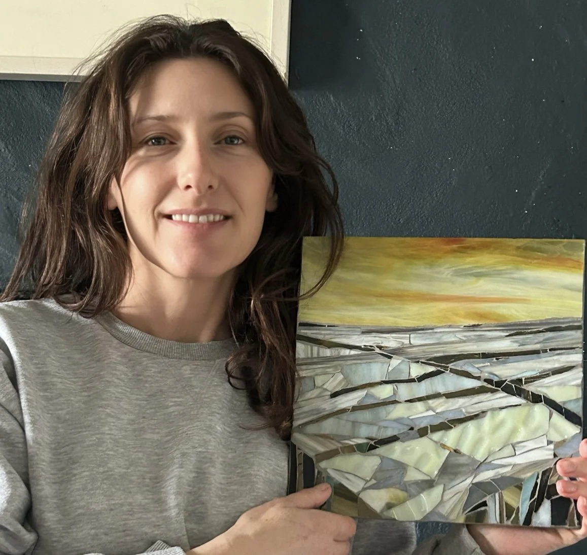 Woman smiling and holding a mirror mosaic artwork depicting a landscape with a colorful sky and rolling hills.