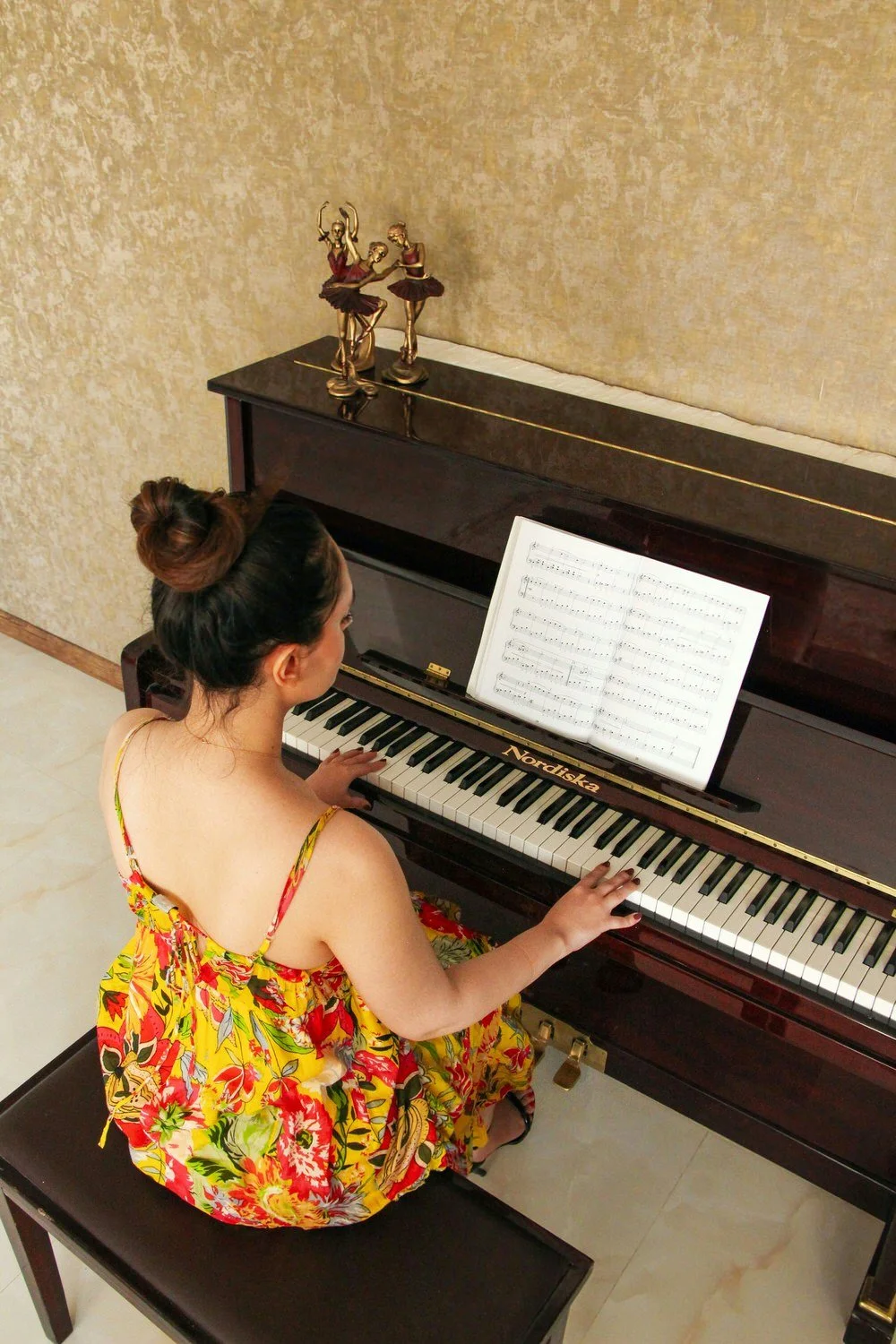 A young woman in a floral dress playing a dark brown upright piano with sheet music on the music stand.