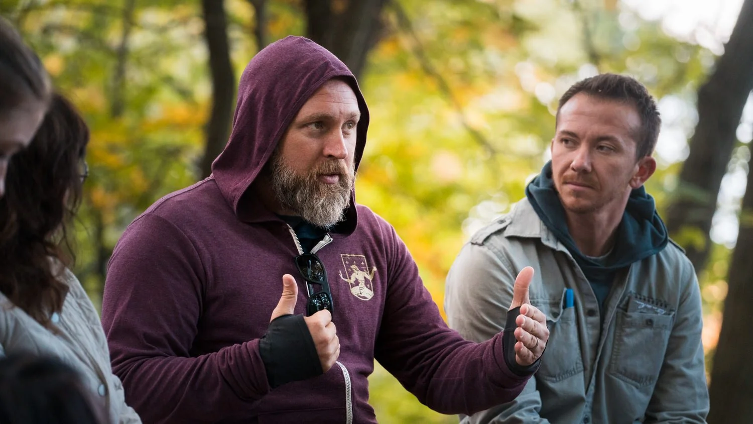 Three people sitting outdoors in a wooded area during fall, having a conversation. The man in the center has a beard, wears a purple hoodie with sunglasses hanging on the neckline, and is gesturing with his hands. The man to his right has short hair, is clean-shaven, and is wearing a gray jacket with a hoodie underneath, listening attentively. The person on the left is partially visible.