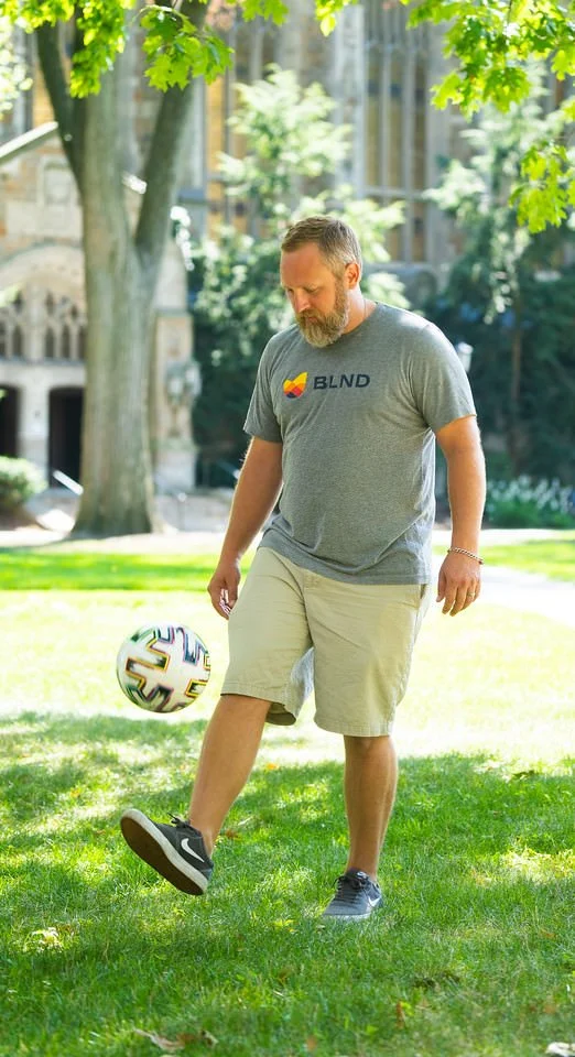 A man with a beard wearing a gray T-shirt and khaki shorts is playing with a soccer ball on a grassy park. Trees and buildings are in the background.