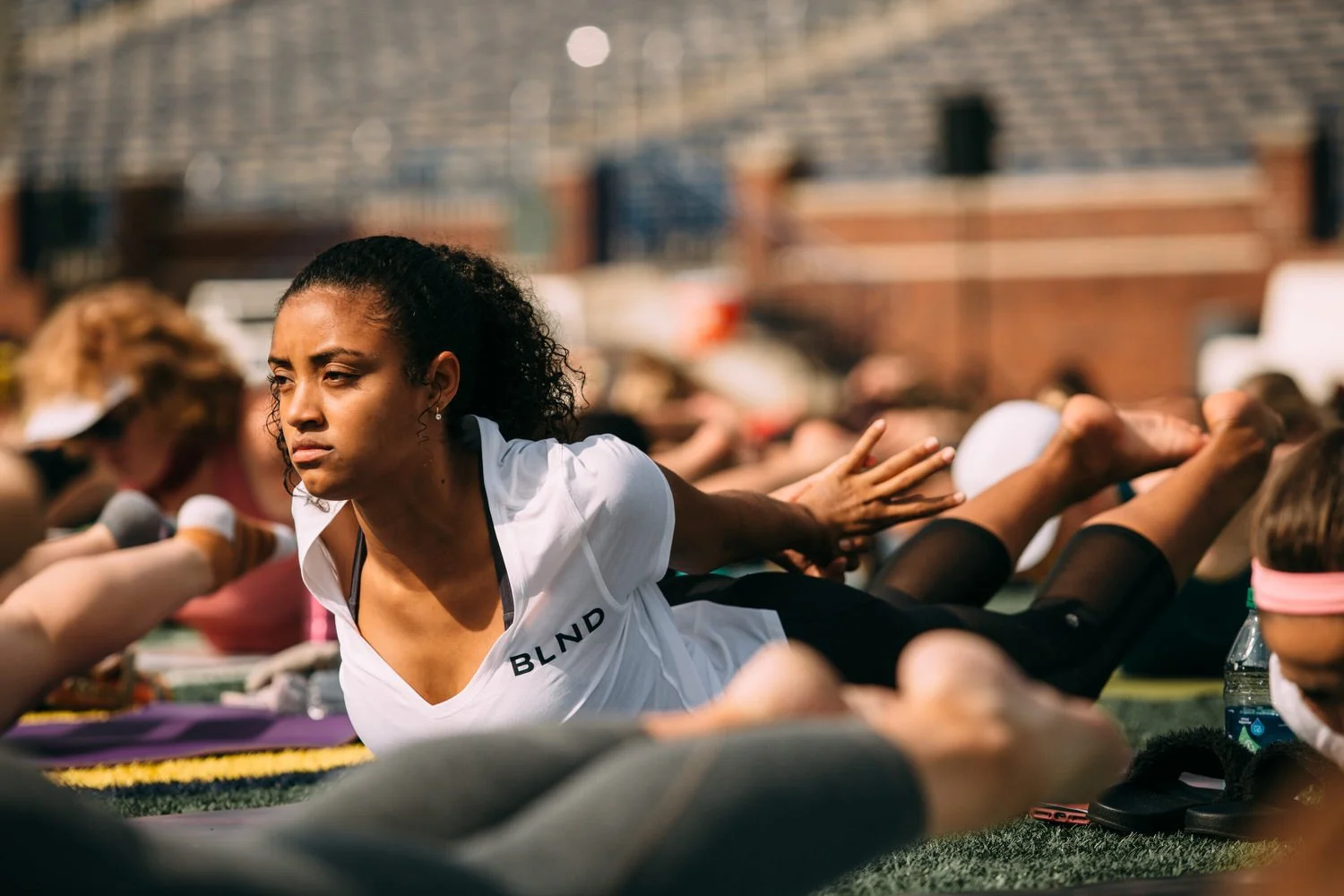 A woman participating in an outdoor yoga class, lying on her stomach with her hands extended forward, wearing a white shirt with "BLND" on it, and surrounded by other yoga practitioners.
